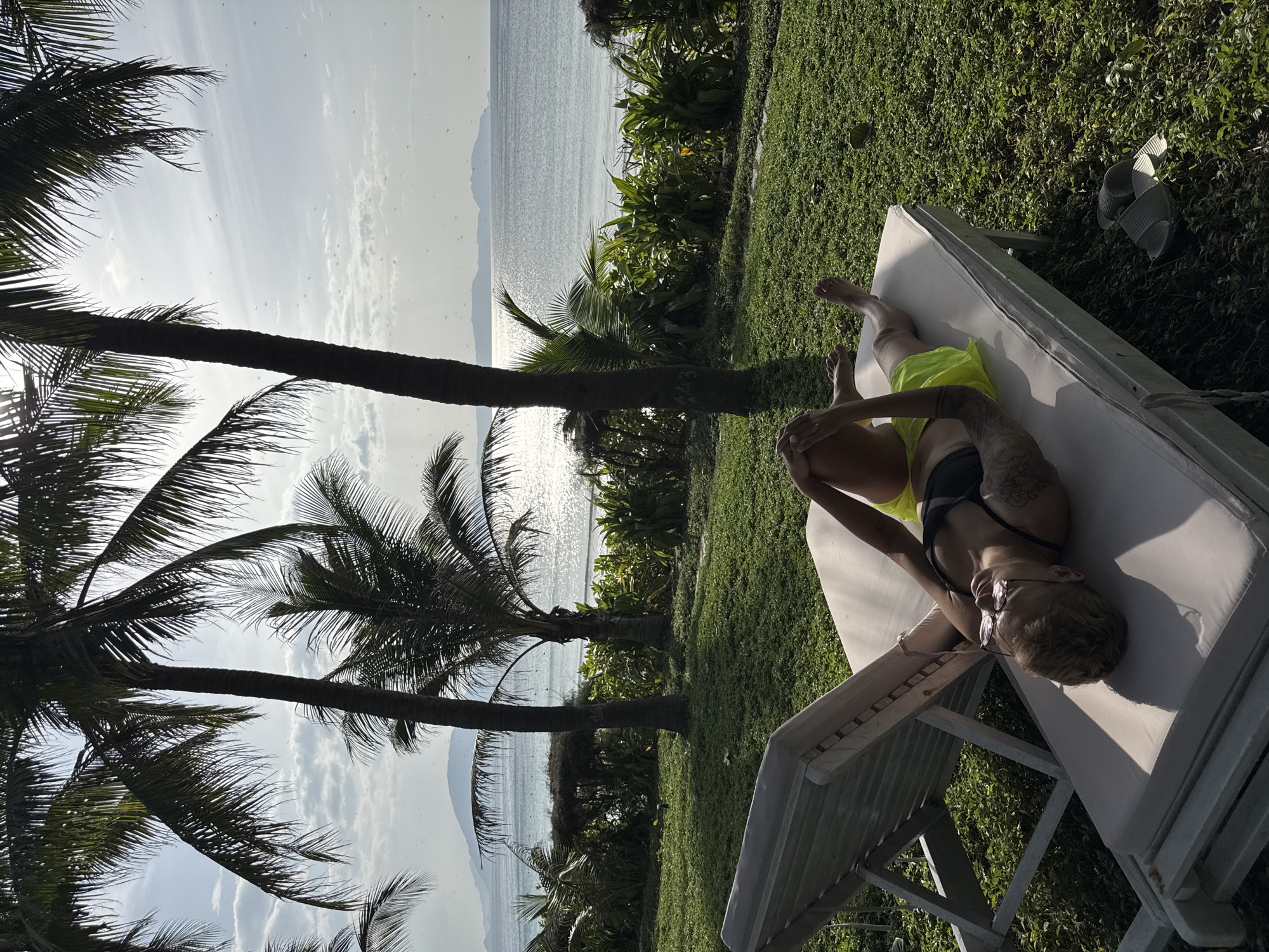 A person with short blond hair and tattoos is lying on a lounge chair on a grassy area overlooking a body of water, likely the sea. The person is wearing sunglasses and a black bikini top with bright yellow shorts. They are holding their hands together in their lap and their legs are bent. Behind the lounge chair are two palm trees, and beyond that is a lush green hedge with more palm trees and foliage. The sea is visible in the background, with the sun reflecting off the water. Mountains are visible in the distance across the water, suggesting a coastal location. The sky is bright with scattered clouds. The scene appears to be in Đông Ninh Hòa, Vietnam.