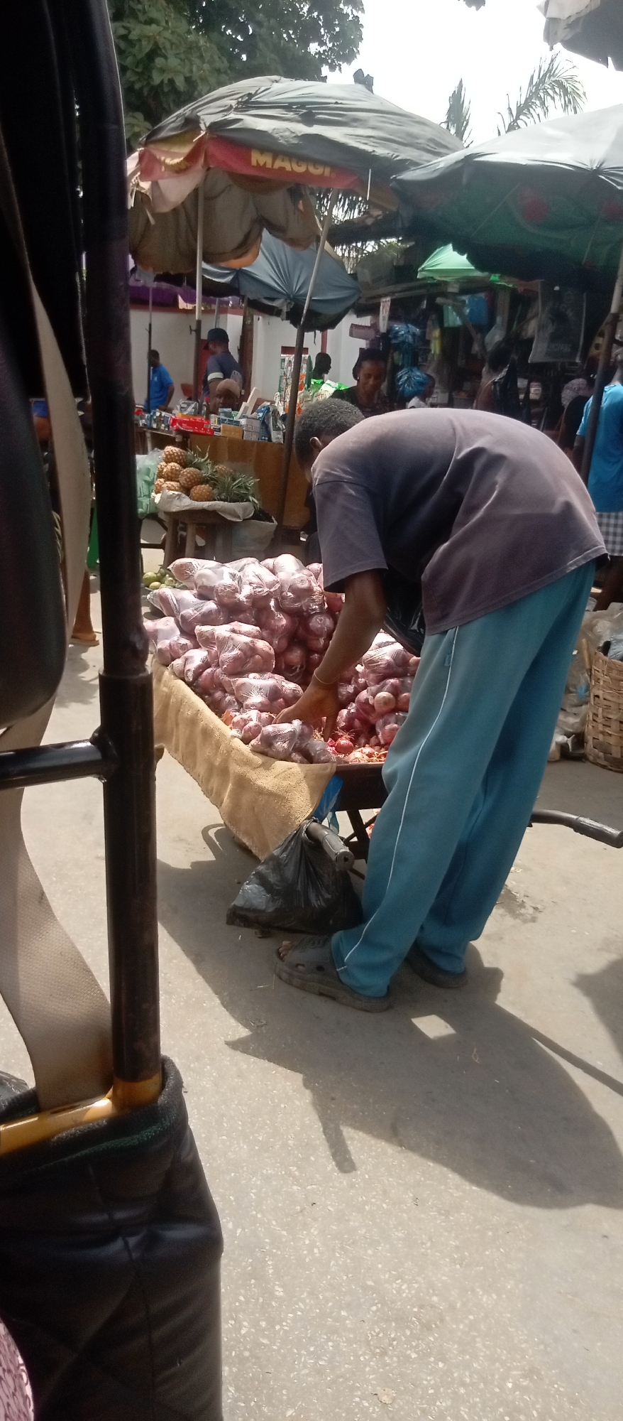 This image captures a vibrant outdoor market scene, likely in Onitsha, Nigeria, under bright daylight conditions. In the foreground, a man, wearing a dark short-sleeved t-shirt and light blue sweatpants with a white stripe, is bent over, arranging or selecting items from a large display of produce. He wears grey slip-on sandals.
The primary produce on display is a substantial pile of red onions, with many individually packaged in clear plastic nets or bags, while others are loose. These are arranged on a cart or stand covered with a light brown, coarse fabric. A black plastic bag hangs from a part of the cart. To the left, part of a vehicle, possibly a tricycle (keke napep), with a black padded frame and a beige strap, is visible.
In the mid-ground, behind the onion stall, another vendor's display includes a pile of fresh pineapples on a white cloth, positioned on a wooden stand. Further into the background, other market stalls with various packaged and unpackaged goods are visible, and several people are present, browsing or working. A distinct white wall can be seen behind these stalls.
Overhead, large market umbrellas provide shade from the sun. One prominent umbrella is dark-colored and clearly displays the "MAGGI" logo in red and white lettering. Another umbrella, partially visible, features a green pattern. Green foliage from trees extends into the top of the frame. The ground is a light-colored paved or concrete surface, with strong, dark shadows cast by the people and market structures, indicating sunny weather. The overall scene depicts a busy, open-air commercial environment.