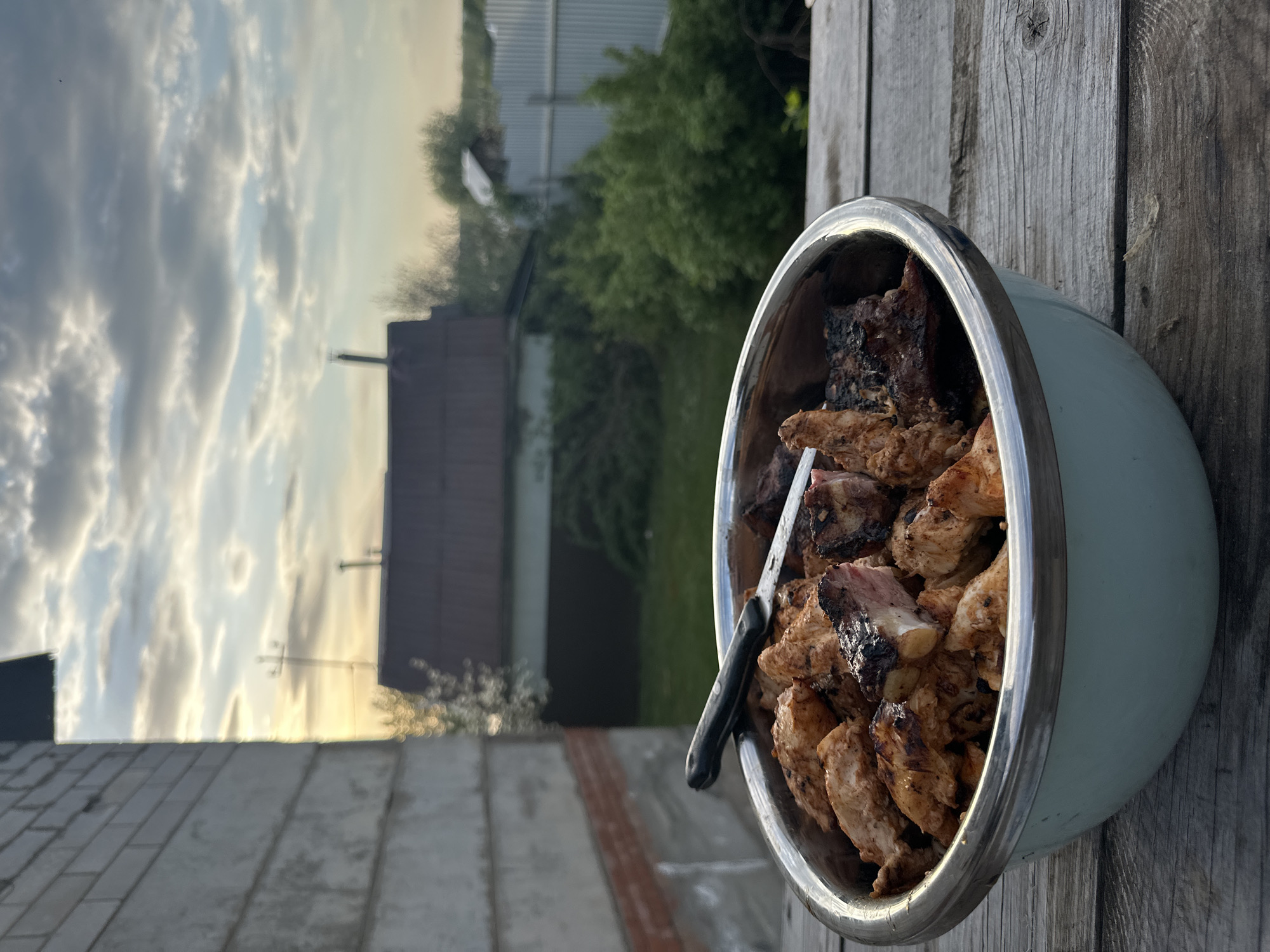 A medium-shot captures a blue metal bowl filled with grilled meat, including some bone-in pieces, alongside a black-handled knife resting within the bowl. The bowl is positioned on a weathered wooden surface, presumably a table.
In the blurred background, glimpses of residential buildings, including a brick structure with visible brickwork, a small tree, and a fence can be observed. The sky is partially visible, displaying a mix of cloudy and clear sections, suggesting either dawn or dusk.
The photograph was taken outside, likely in a backyard or garden setting, within the city of Chaikovsky, Russia. No people are visible in the image.