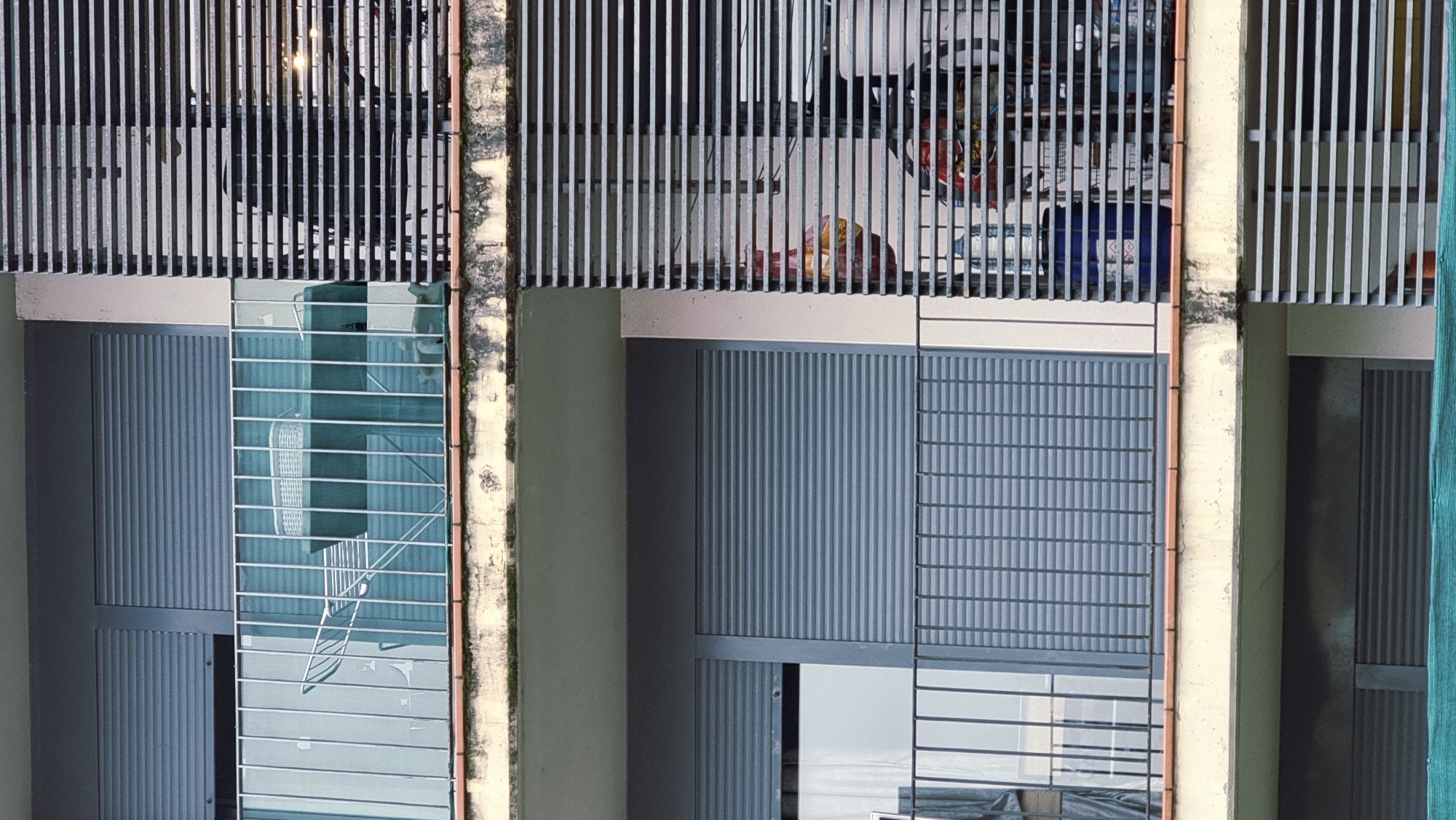 The image shows the exterior of an apartment building in Sant Joan Les Fonts, Spain. The building has multiple balconies with gray metal railings and shutters on the windows. The balcony on the upper level contains a drying rack with a white basket on it and a green table. The balcony on the lower level has a blue plastic container and some other items that are partially obscured by a slatted metal screen. The building appears to be constructed of concrete and has some visible signs of wear and tear. The overall scene is residential and appears to be an ordinary day.