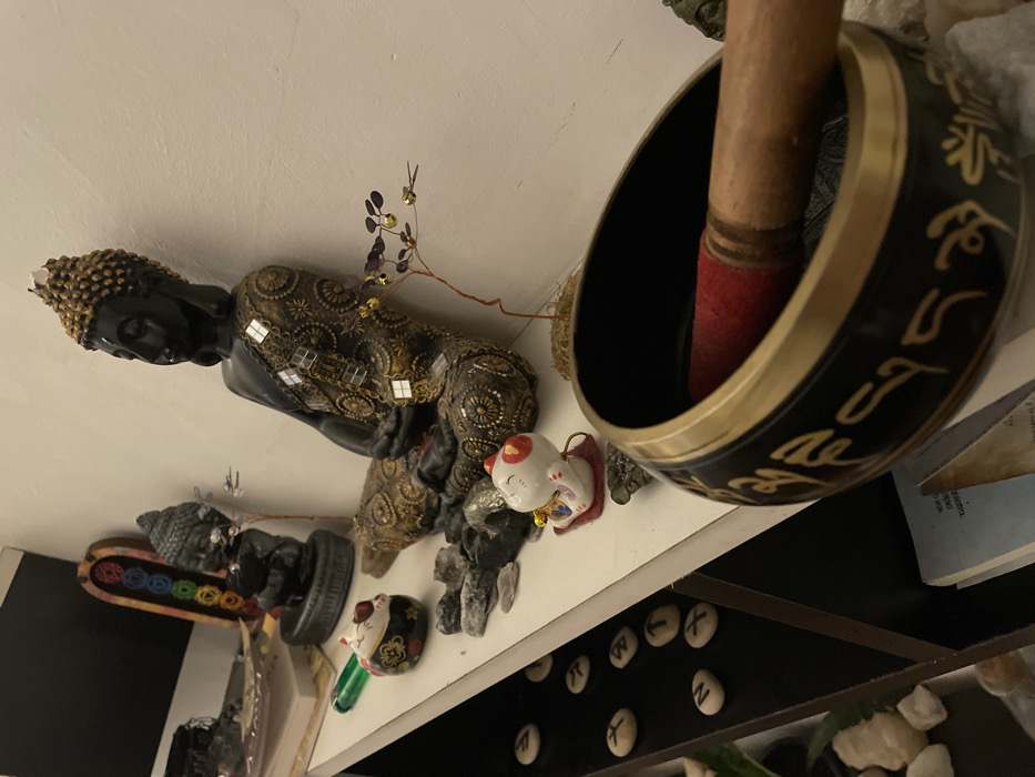 A shelf displays spiritual artifacts. A large Buddha statue with gold accents sits to the left, alongside a smaller Buddha on a rainbow tray. A maneki-neko cat figurine is positioned near rock formations. A singing bowl with a wooden mallet dominates the right side of the frame. Beneath the shelf, rune stones are visible.