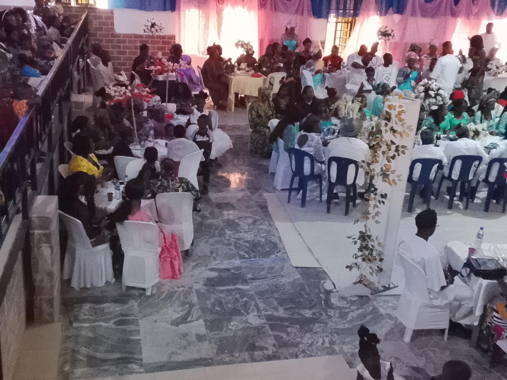 The image depicts an indoor gathering of people seated at tables in a large hall. Most of the people are dark-skinned, some dressed in traditional African clothing, while others are in Western attire. Some are seated at tables adorned with floral arrangements. There is a balcony area with additional seating. The hall is decorated with blue and pink drapes. Based on the reference information, it is likely that this scene is located in Jos, Nigeria.