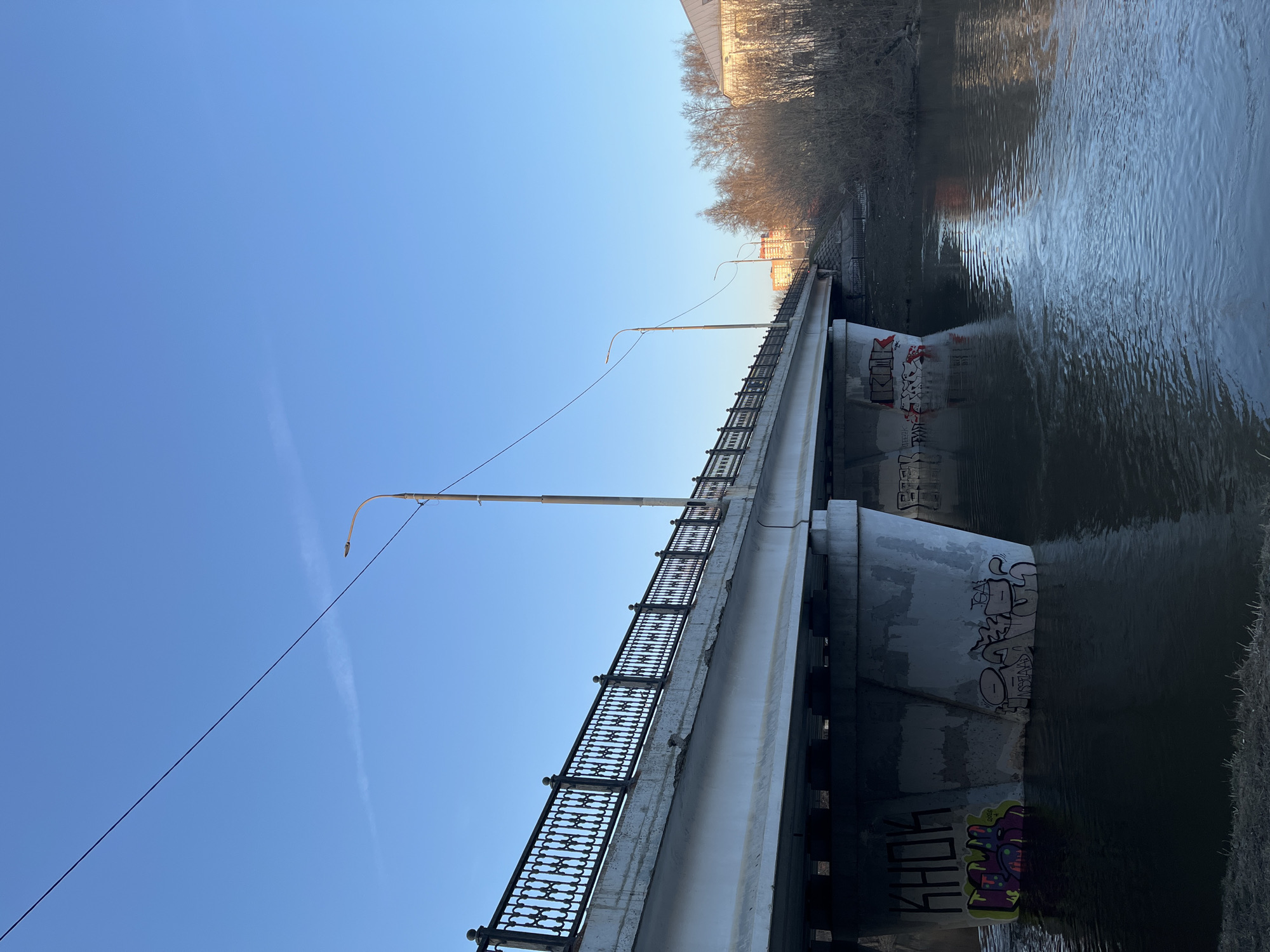 The image depicts a bridge over a river in the city of Podolsk, Russia. The bridge is made of concrete and steel, with a black metal railing along its sides. Graffiti is visible on the bridge supports. The river flows underneath the bridge. The sky is clear and blue. Trees and buildings are visible in the background. A utility pole is also visible above the bridge.