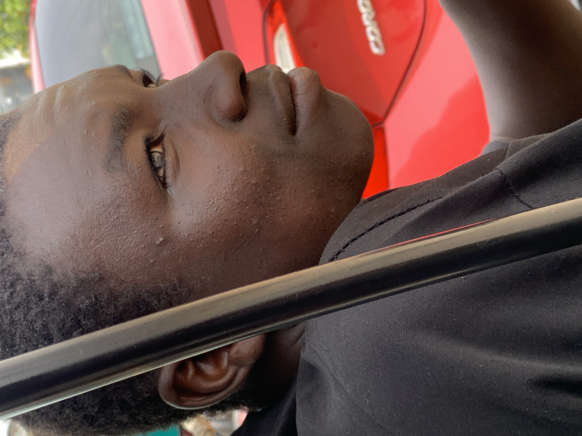 A close-up, angled photograph shows a dark-skinned man with short, curly hair, positioned near a red car identified by the "Corolla" emblem. The man is wearing a dark-colored shirt. A thin, black rod is positioned horizontally across the middle of the frame, partially obstructing the view of his face. The man's face is partially turned upward. The location is identified as Jos, Nigeria.