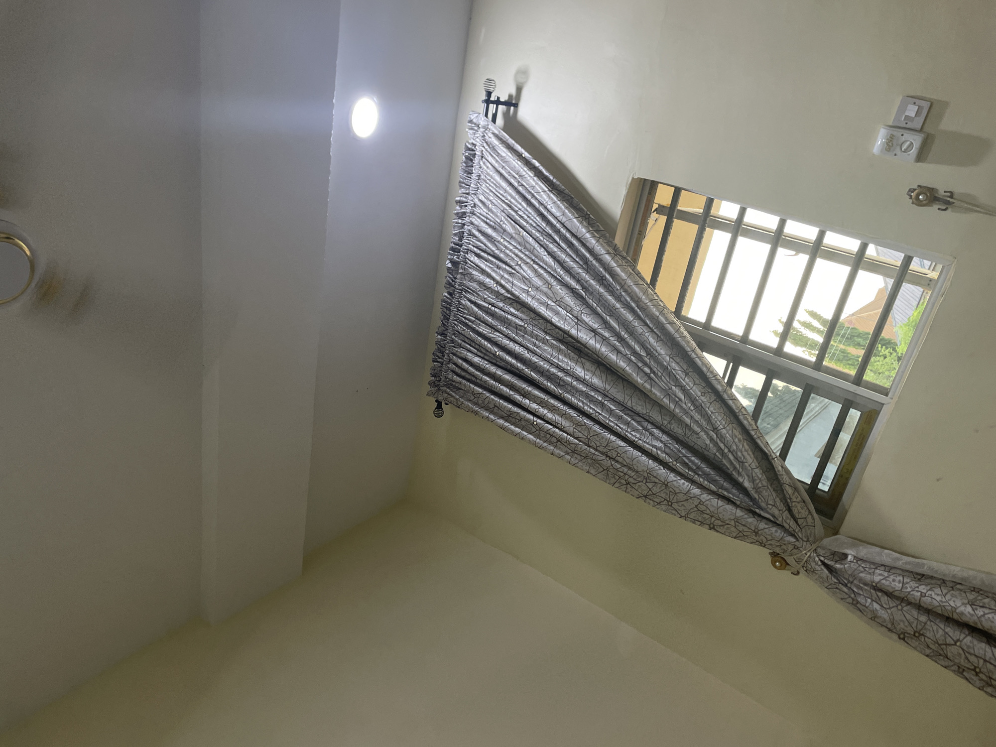 The image depicts an interior view of a room, likely a bedroom, with a window and a curtain. The walls are painted a light yellow, and the ceiling is white with recessed lighting. A decorative curtain with a geometric pattern is partially drawn over the window, revealing a glimpse of the outdoors. The window has horizontal bars for security. On the wall near the window, there are two electrical outlets and a light switch. A curtain tie-back, a metal hook, and a coiled cord are also visible on the wall. The scene is illuminated by natural light coming through the window and artificial light from the ceiling fixture. This image appears to have been taken in Howayi, Nigeria.