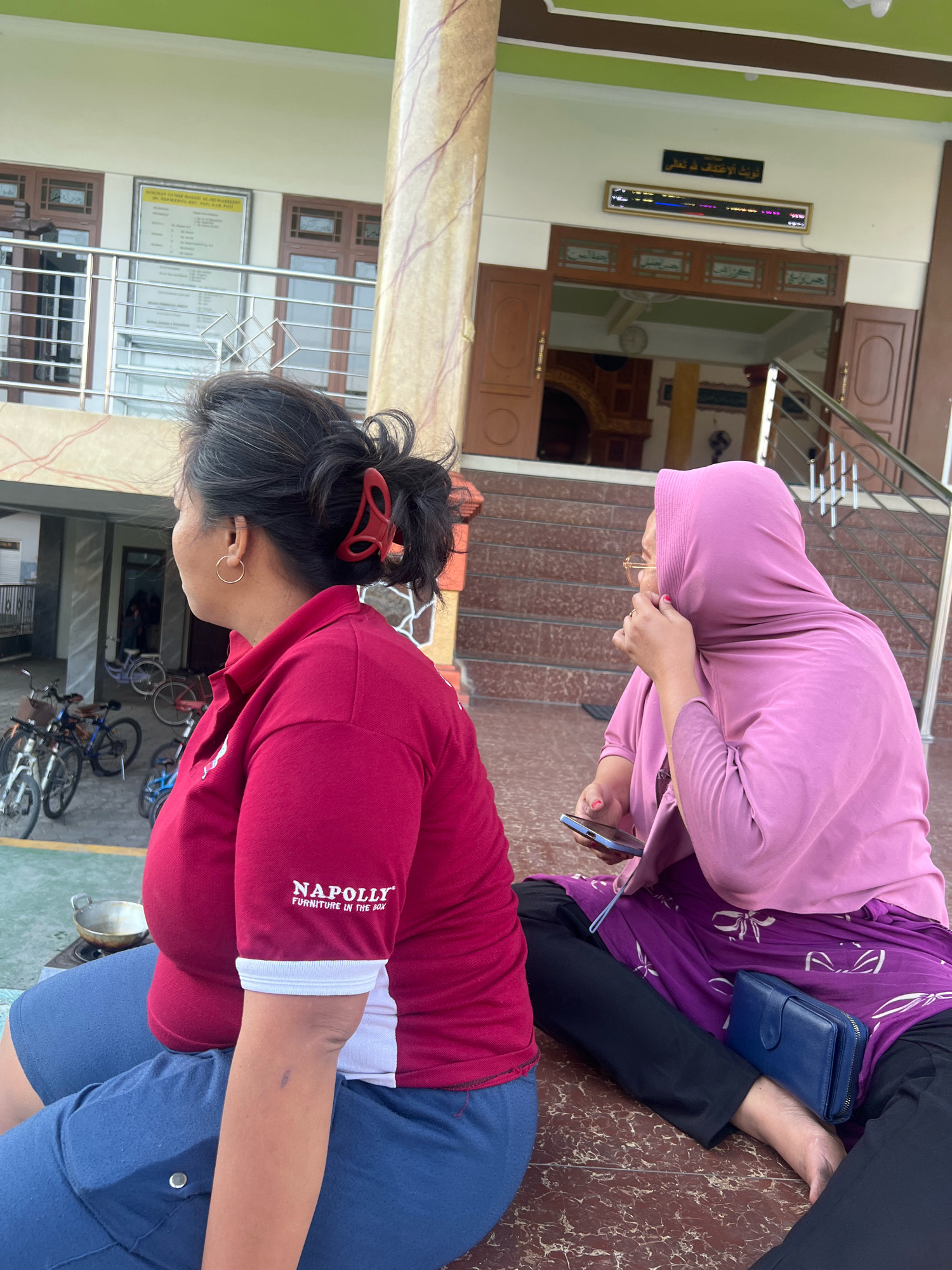 The image shows two women sitting on the ground outside a building. The woman on the left is wearing a red shirt with "NAPOLLY FURNITURE IN THE BOX" printed on it and blue shorts. She has her hair pulled back with a red clip and is wearing a hoop earring. The woman on the right is wearing a pink hijab, glasses, and a purple skirt over black pants. She is holding a smartphone and has a blue wallet on her lap. In the background, there is a building with a green roof and light-colored walls, featuring wooden doors and windows. There are bicycles parked to the left of the building. The overall setting appears to be in an outdoor area of a community space, potentially a religious or educational institution, given the Arabic inscriptions above the entrance. The scene suggests a casual moment of rest or waiting. This location is identified as Kecamatan Pati, Indonesia.