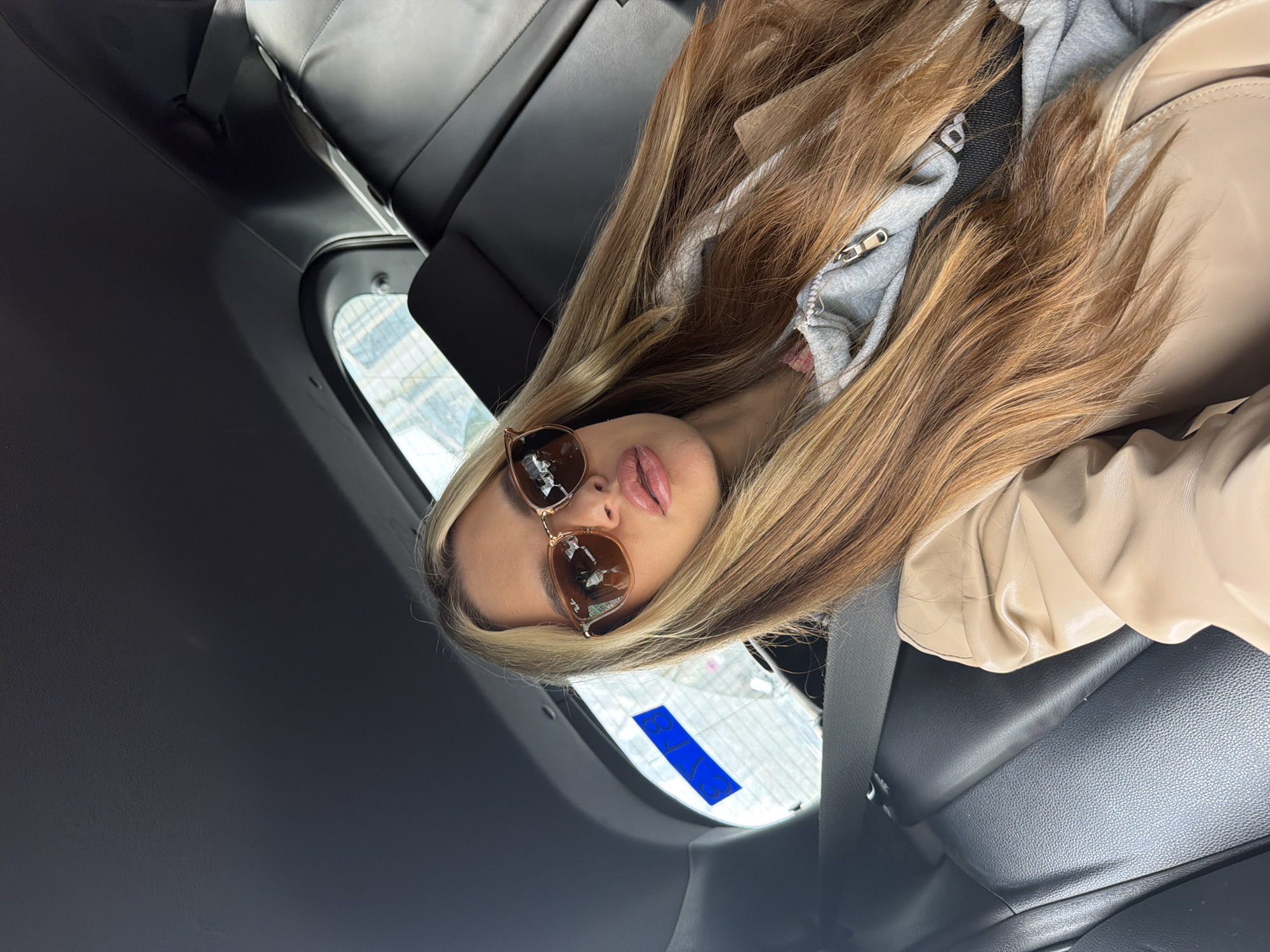 A female individual is seated in the rear passenger compartment of a vehicle, angled toward the camera for a selfie. She has long, straight, multi-toned blonde and light brown hair. Her facial features include full lips coated with light pink lipstick, and she wears hexagonal Ray-Ban sunglasses with light brown frames. Her attire consists of a light grey zipped hoodie, a pink garment underneath, and a tan or beige faux leather jacket. A black strap, possibly from a bag, crosses her chest. The car's interior features dark upholstery, including black seats and headliner, with a visible dark grey seatbelt. Through the rear passenger window, a modern building with a grid-patterned glass facade is visible. A blue rectangular sign or reflection displaying the white characters "B 78" is partially discernible against the building backdrop. This "B" identifier strongly indicates the location is in Berlin, Germany.