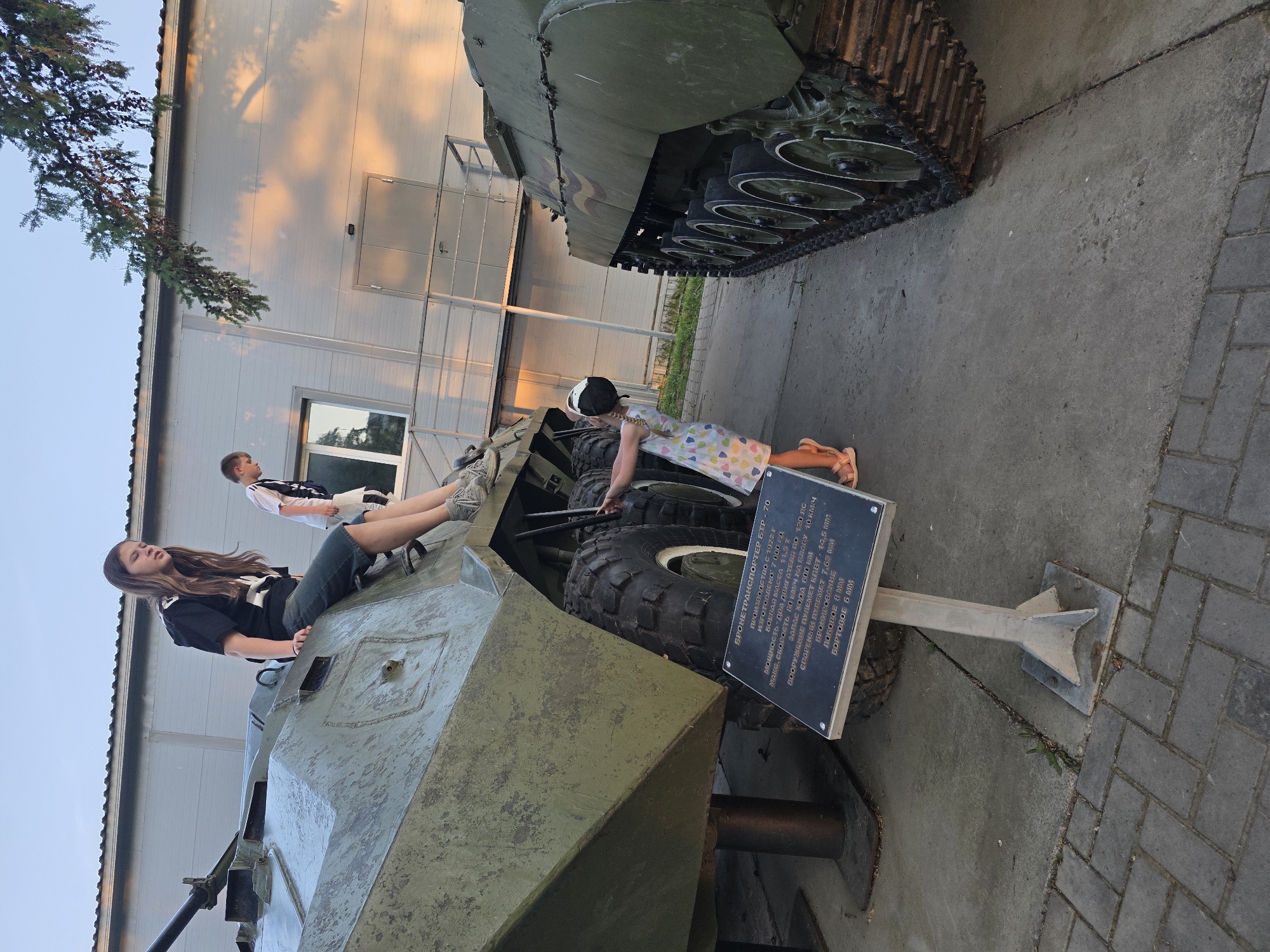 The image shows two children climbing on military vehicles in what appears to be an outdoor exhibit. One child, a girl with long brown hair, sits on the front of a green armored vehicle, while another child, a girl in a polka dot dress and baseball cap, stands near the back tires of the same vehicle. A third child is visible standing near the vehicle. Adjacent to the vehicle is a sign with text in Russian, identifying the vehicle as a BTR-70 armored personnel carrier, produced from 1972 onward. In the background, there is a larger, tank-like vehicle. The scene takes place outdoors, with a building visible in the background. The location of the city of Votkinsk, Russia is NOT discernable from the image.