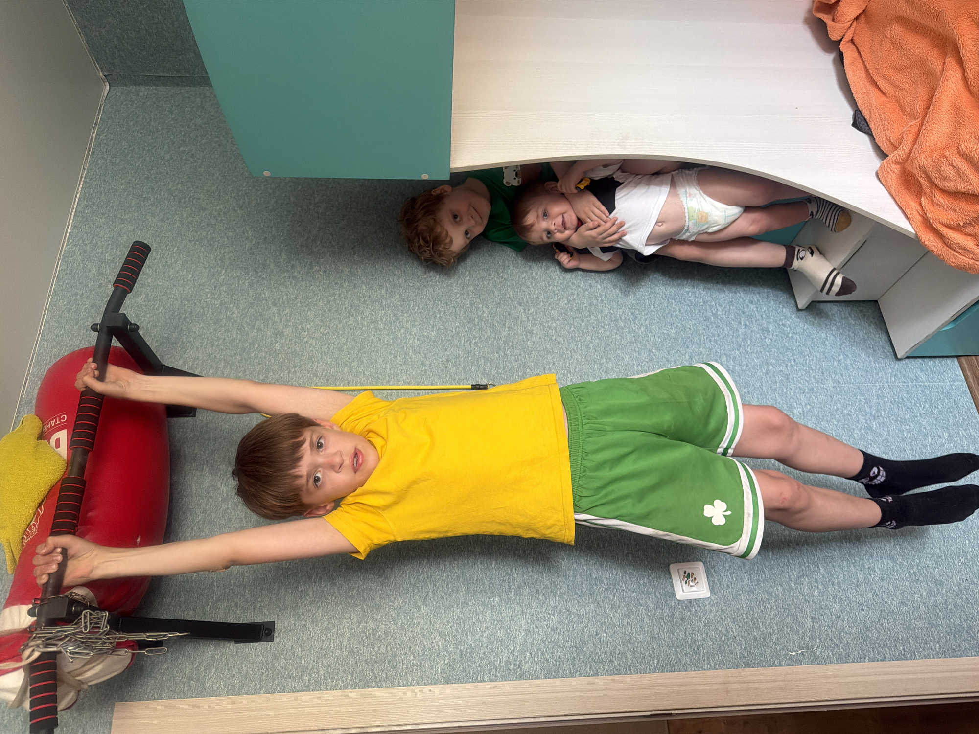 The image depicts an interior space with three young children. A boy wearing a yellow t-shirt and green shorts with a white shamrock design is lying on the floor, arms outstretched and holding onto a red punching bag apparatus. Another boy wearing a green t-shirt is positioned partially under a white and teal-colored structure, possibly a bed or cabinet. A third, smaller child wearing a diaper, black and white t-shirt, and patterned socks lies near the second boy, also partially under the structure. An orange towel is seen on top of the structure. The floor is covered in a light blue speckled linoleum. A yellow resistance band is visible near the boy doing the pullups. A small white square is visible near the boy's legs. No information available about the location, Noviy, Russia.