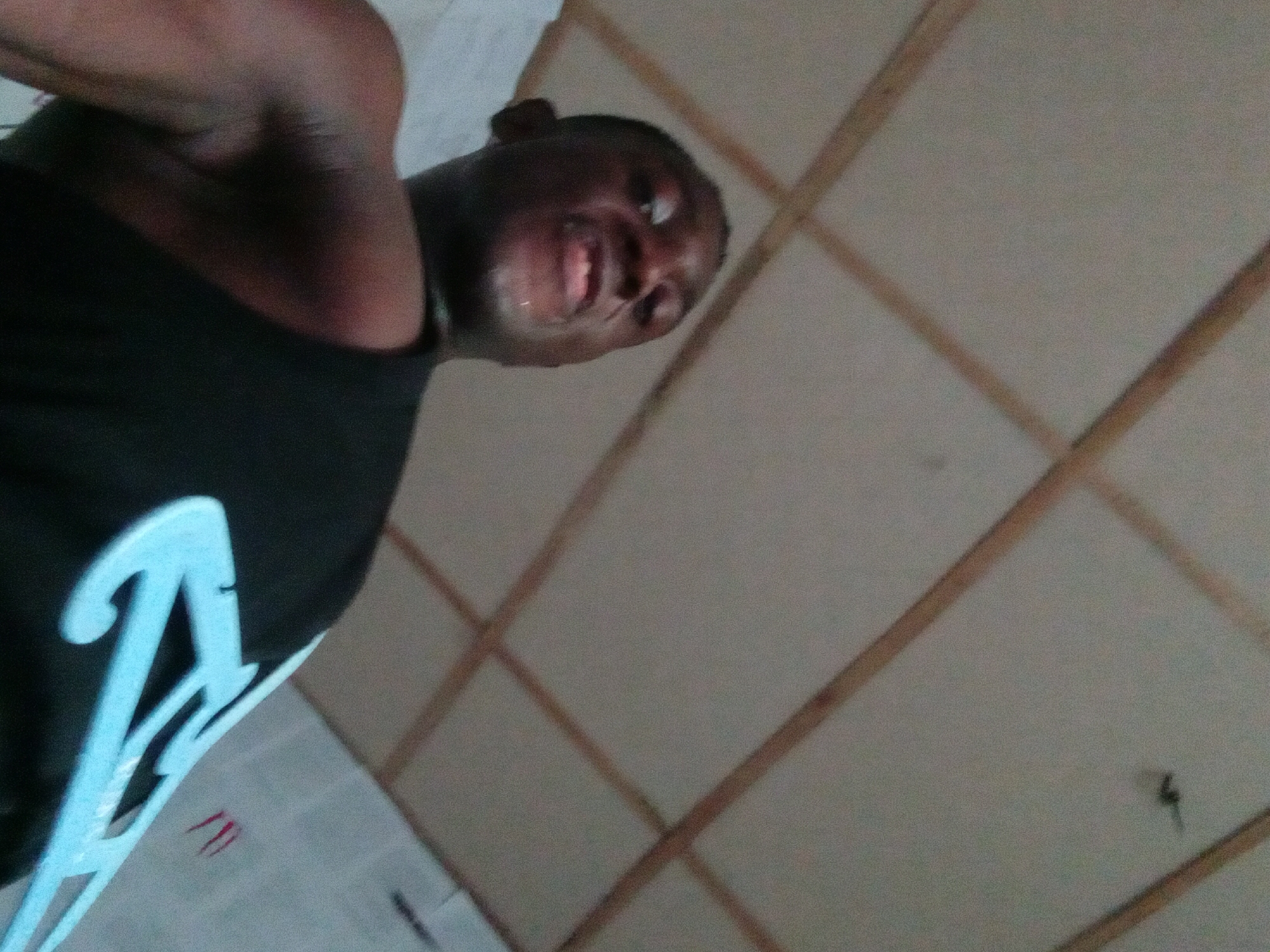A person with dark skin is wearing a black tank top with a light blue design. The person is looking up at the camera and smiling. The background is a ceiling with a grid pattern. The location is identified as Akwete, Nigeria. No further details are available.