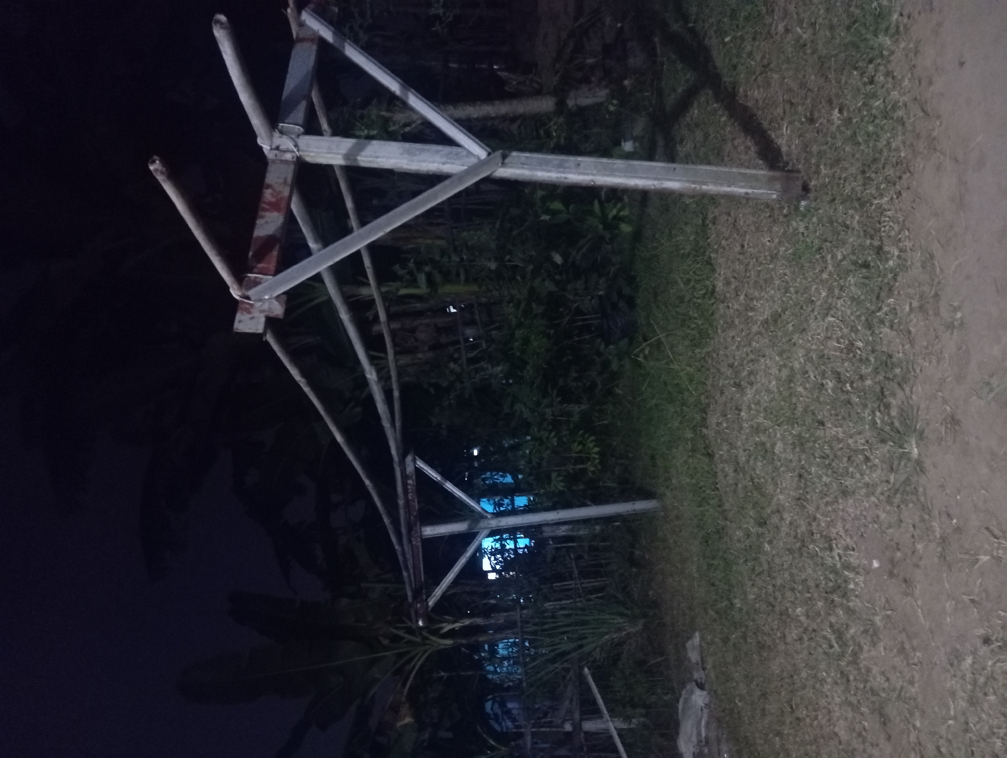 The image depicts an outdoor scene at night, likely in Kutasari, Indonesia, focusing on a weathered metal drying rack. The rack, constructed from galvanized metal pipes and angles, stands prominently in the center, showing signs of rust and age. It is supported by two main vertical posts, each reinforced by diagonal braces forming a 'V' shape. Horizontal bars extend from the top of the posts, creating a structure for hanging laundry.
The surrounding environment is somewhat overgrown and appears to be a residential backyard or a communal outdoor space. Lush green vegetation, including what appear to be banana plants or similar large-leafed tropical flora, forms a dense backdrop behind the drying rack. A low fence, possibly made of bamboo or wood, is partially visible behind the vegetation, and some bright blue light emanates from beyond it, suggesting a lit building or structure. The ground is covered with dry grass and dirt. The overall lighting is dim, characteristic of nighttime, with some shadows cast by the drying rack and surrounding elements. There are no people or discernible actions taking place in the image.