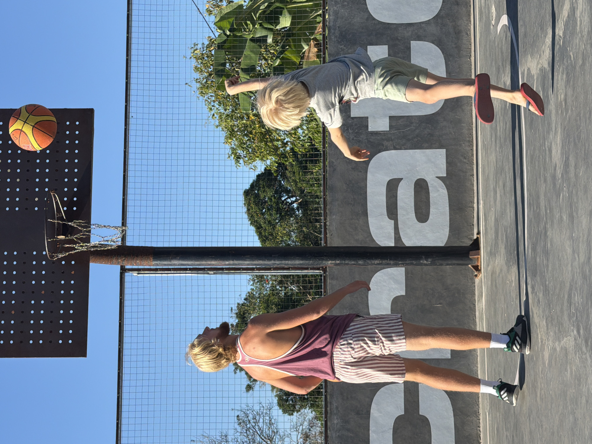Two people are playing basketball on an outdoor court in Mengwi, Indonesia. A man with blonde hair and a beard, wearing a burgundy tank top and striped shorts, stands on the left, looking up at the basketball hoop. On the right, a child with blonde hair, wearing a grey t-shirt and light green shorts, is in mid-air, attempting a shot. The basketball is visible in the air, near the hoop. The court has a concrete surface with painted lines, and a grey wall with large white letters is visible in the background. A black metal fence encloses the court, and trees and foliage are visible beyond the fence. The sky is clear and blue.