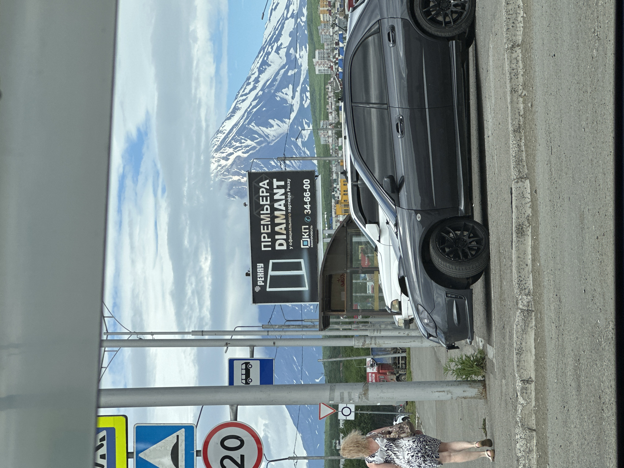 The image depicts a street scene in Petropavlovsk-Kamchatsky, Russia, with a prominent snow-capped volcano in the background. In the foreground, a woman with blonde hair, wearing a black and white patterned dress and carrying a handbag, walks away from the camera on a sidewalk. She is wearing sandals. Adjacent to the sidewalk are several traffic signs, including a speed limit sign indicating "20", a warning sign for a speed bump, and a bus stop sign. Two cars are parked on the street: a dark gray sedan in the foreground and a white SUV behind it. A large billboard advertising "RENAY DIAMANT" is visible, along with a bus shelter and buildings in the middle ground. The sky is partly cloudy, and the overall environment suggests an urban setting near a natural landscape.