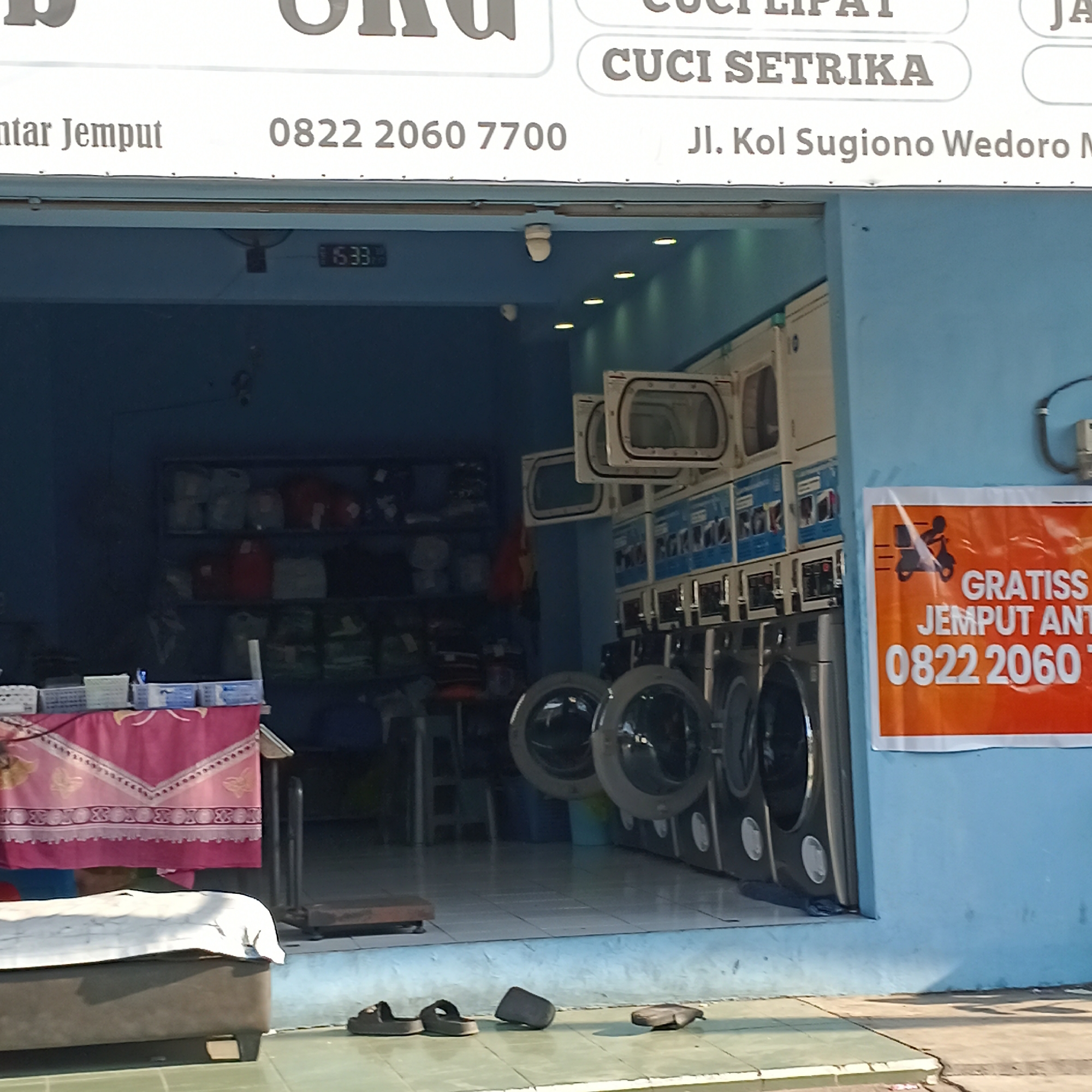 The image shows the exterior of a laundromat in Wedoro, Indonesia. The sign above the entrance reads "CUCI LIPAT" and "CUCI SETRIKA", indicating laundry and ironing services. Below this, the address "Jl. Kol Sugiono Wedoro M" is visible, along with phone numbers "0822 2060 7700" and "0822 2060 7".
Inside the laundromat, multiple washing machines are stacked vertically and horizontally. Some washing machine doors are open, revealing their drums. A digital clock on the ceiling displays "15:33". On a shelf inside, various bags of laundry are stored.
In front of the laundromat, a table is covered with a pink patterned cloth, and various items are placed on it. A dark-colored sofa or bench is positioned in the foreground, with a white cloth draped over it. A pair of sandals is placed on the ground to the right of the sofa, and another dark object, possibly a bag or shoe, is near the entrance. An orange sign on the blue wall prominently displays "GRATISS JEMPUT ANT" and the phone number "0822 2060 7". The ground in front of the laundromat is tiled with a lighter green and grey pattern.