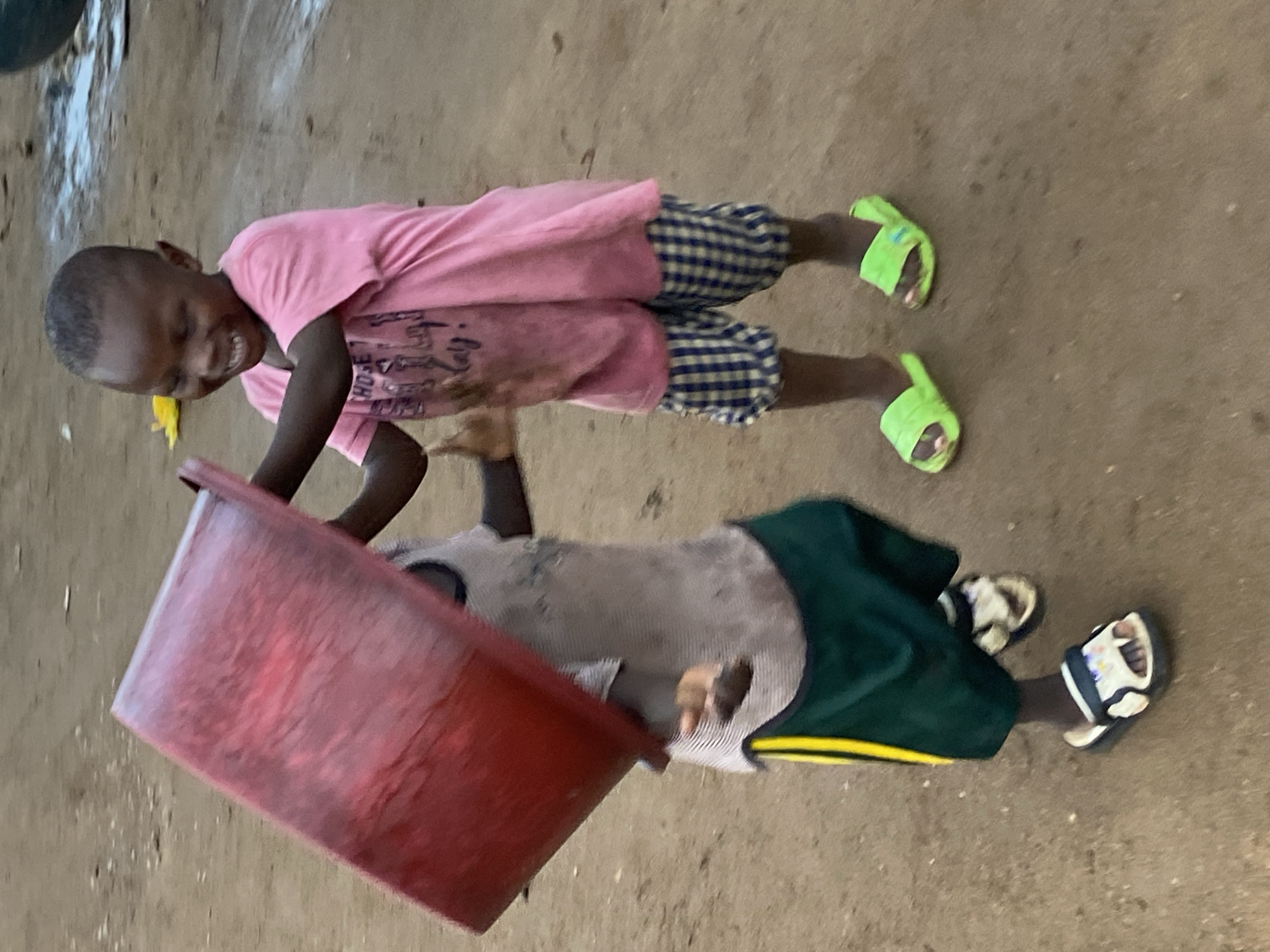Two young boys are engaged in play in a dusty, outdoor environment, possibly a street or yard in Bukuru, Nigeria. The boy on the right, wearing a pink t-shirt and blue and yellow checkered shorts, is laughing and holding a large red plastic basin. The boy on the left, in a grey and white striped t-shirt and green shorts with a yellow stripe, is partially obscured by the basin, but appears to be interacting with it. Both boys are wearing sandals. The ground is composed of dirt and sand. The lighting suggests it may be late afternoon or early evening.