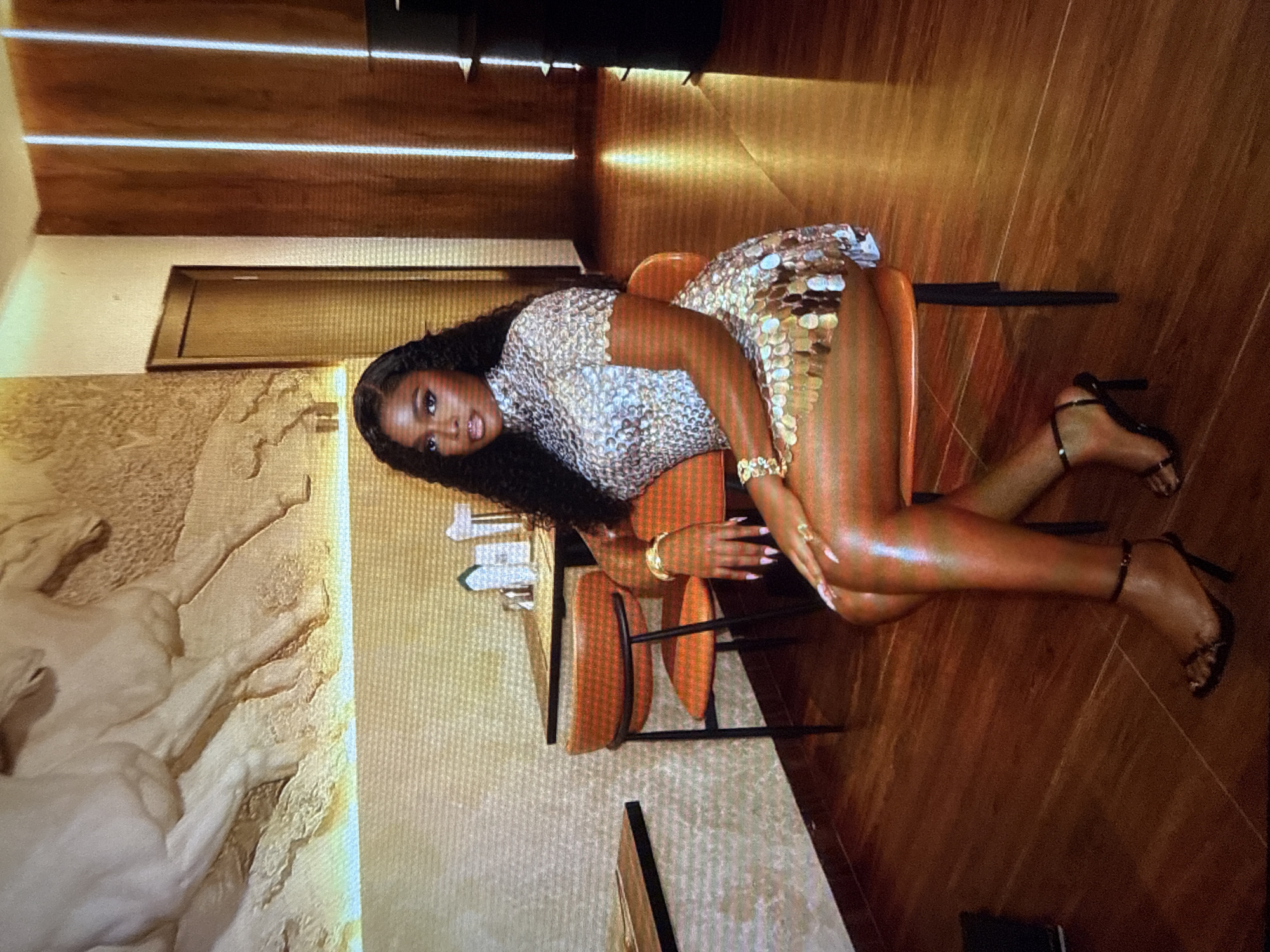 A Black woman is seated on an orange chair in what appears to be a restaurant or lounge, with wooden floors and textured walls. The wall behind her features a relief sculpture of horses. The woman has long, dark, curly hair and is wearing a short, silver, sequined dress and black high-heeled sandals. She is adorned with several gold bracelets and rings. Her legs and arms have a shimmering sheen. The lighting in the establishment is warm and ambient, with vertical light fixtures visible in the background. The tables and chairs suggest a dining or social setting. The specific location is identified as Abuja, Nigeria.