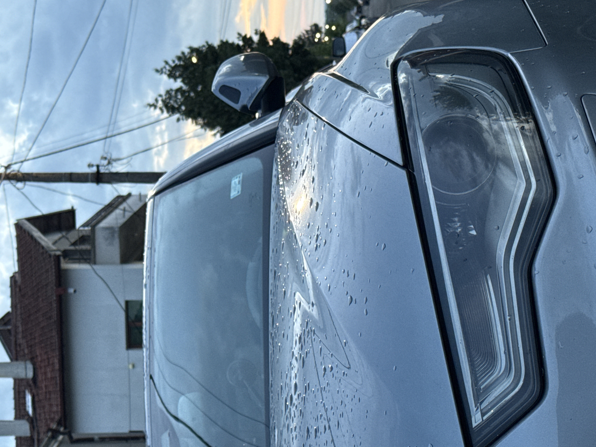 A close-up, slightly angled view of the front passenger side of a light gray car, possibly in the city of Burgas, Bulgaria. The car is wet, displaying numerous water droplets on its hood and headlight. Visible features include the side mirror, windshield, and the intricate design of the headlight. The car is parked near a building with a tiled roof and white walls. Overhead, power lines traverse the sky, with a tree visible in the background. The sky suggests either sunrise or sunset with a muted color palette.