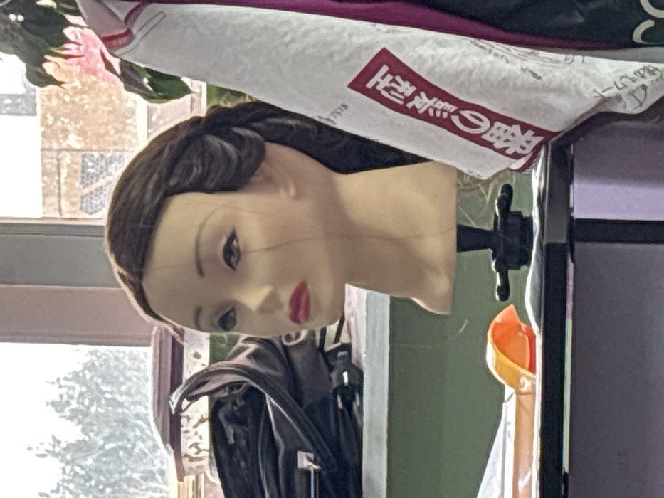 A beige female mannequin head, featuring a dark brown wig styled with voluminous waves, is centrally positioned, facing left in profile. Its painted facial features include dark eyebrows, black winged eyeliner, and red lipstick; two thin strands of hair are visible descending vertically on its left cheek. The mannequin head is mounted on a black stand, resting on a dark-colored, flat surface, likely a desk. To its immediate left on this surface, an orange U-shaped object is present. Further left and slightly behind, an open black bag is visible on a lighter shelf. To the right of the mannequin, a white cloth or towel with vertical red Japanese characters, translating to "Traditional Manufacturing Method," hangs, partially obscuring a dark purple garment. The background reveals a large window overlooking blurred outdoor scenery, featuring evergreen trees to the left and an urban building with a distinct lattice pattern to the right. Indoors, a green plant with pink blooms is visible behind the mannequin's right side. The precise location within the city of Chaikovsky, Russia, cannot be determined from the visual information provided.