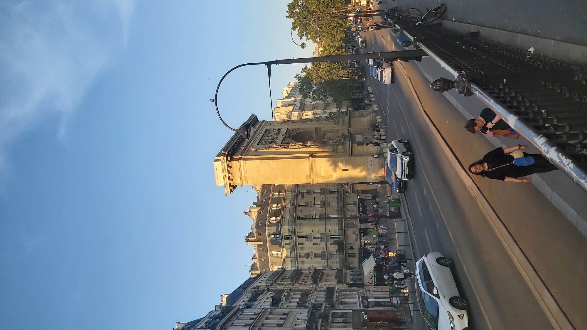 The image captures a street scene in Paris, France, specifically at the Arc de Triomphe du Carrousel. The sun is setting, casting a warm golden light on the stone structures and buildings. In the foreground, a black metal railing is visible, with two people walking on the sidewalk beside it. The woman in front wears a black t-shirt and pants, carrying a blue bag over her shoulder. The person behind her, wearing a black t-shirt and jeans, is looking down at an orange and blue bag.
Further back, a street with traffic is depicted. Several cars are driving on the road, including white SUVs and a white station wagon. Pedestrians are visible on the sidewalks on the other side of the street, some near the entrance to the Arc de Triomphe du Carrousel, which stands prominently in the mid-ground. The monument is constructed of stone and features intricate carvings. On the left side of the street, buildings with mansard roofs, typical of Parisian architecture, are lined up. Trees with green foliage are present on the right side of the street. The sky is a clear blue with a few wispy clouds, indicating it is late afternoon or early evening.