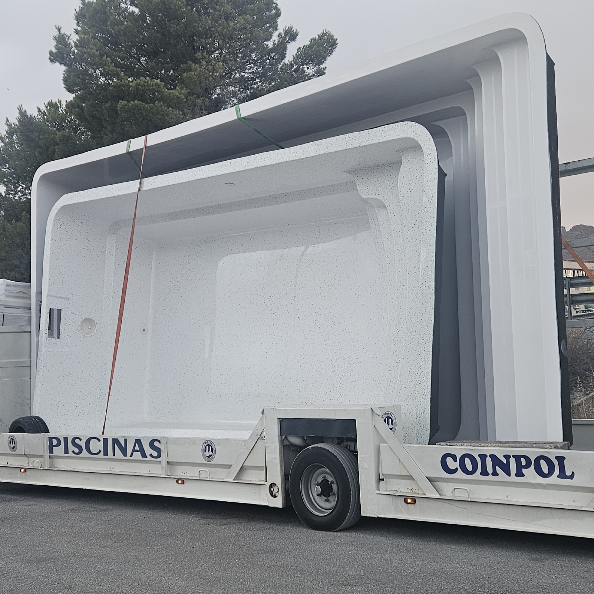 The image captures a trailer transporting a large, white, speckled swimming pool on a road in Albacete, Spain. The pool is white with dark speckles and has a textured interior. It appears to be nested within other similar structures, forming a stack. A red strap is visible securing the pool to the trailer. The trailer itself is white with blue lettering that reads "PISCINAS" on one side and "COINPOL" on the other, along with circular logos. One of the trailer's wheels is visible, resting on asphalt. In the background, trees with green foliage and a slightly overcast sky are present. A sign with the word "AUR...ANT" is partially visible on a structure behind the trailer. The overall scene suggests a transportation or delivery operation.
