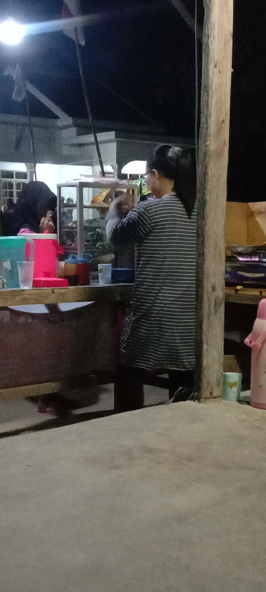 This is a nighttime, medium-shot, vertical image taken at a street-side food stall in Utama Jaya, Mataram, Indonesia. A woman wearing a striped long-sleeved shirt and glasses is standing behind a glass display case, holding something in her hands as if preparing food or drinks. To her left, another person wearing a black hijab is partially visible, also interacting with items in the display case. The stall has a wooden counter with various items including plastic cups, a pink thermos, and condiment bottles. In the background, there is a white building structure and overhead lighting, suggesting an open-air dining or service area. The ground is concrete, and the overall lighting is dim, typical of a nighttime outdoor scene. A large wooden pole is prominent on the right side of the frame.