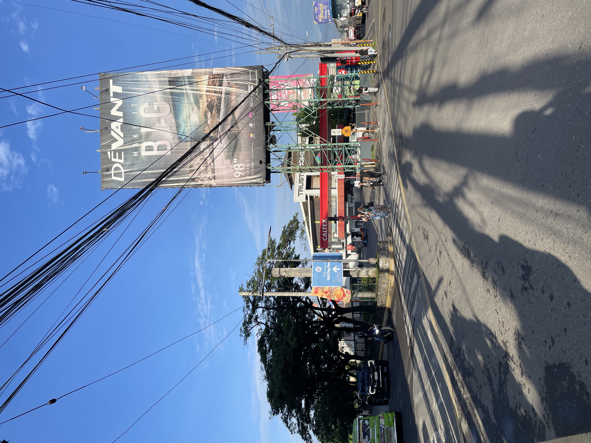 This vertical image captures a bustling street scene in Davao City, Philippines, under a clear, bright blue sky with sparse white clouds. The sun appears to be low in the sky, casting long, distinct shadows of trees and other objects across the asphalt road, suggesting it's either morning or late afternoon.
In the foreground and spanning the left side of the frame, numerous thick utility cables crisscross against the sky and in front of a large billboard. This billboard prominently displays the text "DEVANT MAKE IT BIG" and "98" ULTRAHD" with an abstract, possibly futuristic, image. Below this, a green metal scaffolding structure supports what might be another sign or a part of the advertising infrastructure.
To the right of the street, a Caltex gas station is visible with its red and white branding. Next to it, a building shows the partial text "TRUSTHO". Further down the street, another building or establishment has a blue sign with "PIBILLION". Pedestrians are scattered along the sidewalk and near the buildings, some waiting, others moving. A scooter/motorcycle is parked, and a green jeepney (a common form of public transport in the Philippines) is partially visible in the bottom right corner, alongside a black SUV. In the distance, a bus is also discernable.
A prominent street sign points "This way to SM City Dav," confirming the Davao City location. Large, leafy trees line the street, contributing to the shadows on the road. The street itself features yellow markings and what appears to be a zebra crossing. The scene depicts a typical urban environment with commercial activity and daily commuting.
