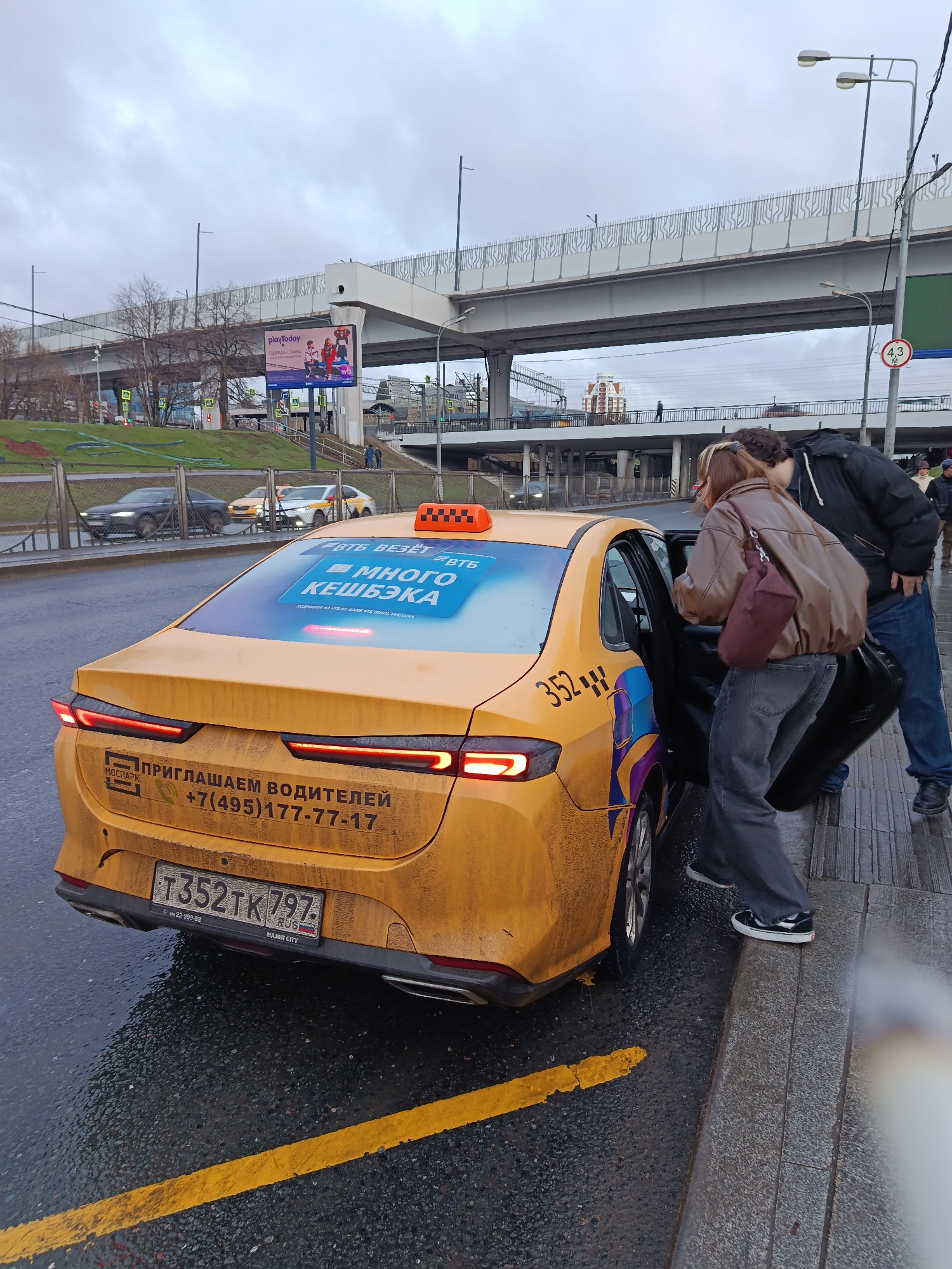 The image captures a yellow taxi with the license plate T352TK 797, parked near a curb in an urban setting. The taxi is emblazoned with advertisements and recruitment information for drivers, including the phone number +7(495)177-77-17. A person is entering the taxi while another stands by the open door. Above the taxi and street level, there is an overpass and elevated roads, suggesting an overpass or highway interchange. Other cars are visible in the background. On the left, a billboard displays the image of "playToday," likely an advertisement for a product or service. The day appears overcast.