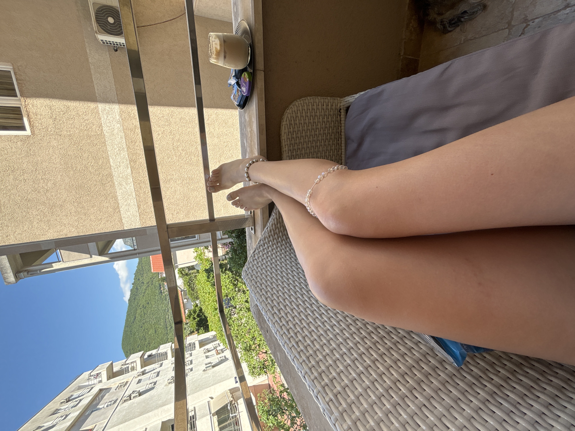 The image presents a first-person perspective from a balcony in Budva, Montenegro, during daylight hours. The lower two-thirds of the frame are occupied by the bare legs and feet of an individual, positioned in a relaxed manner. The skin of the legs is tanned. Each ankle is adorned with a single anklet made of light-colored, possibly pearl or glass, beads. The toenails are painted a light, neutral color. The legs rest upon a light-colored, woven wicker chair or lounger, with a grey fabric cushion visible beneath them.
To the right, on a dark brown balcony ledge, a small, circular silver tray holds a glass containing a layered beverage, consistent with iced coffee topped with foam. Beside the glass, several individually wrapped confectionery items are visible, including one wrapped in purple and others in darker packaging. A metallic horizontal railing extends across the mid-ground of the balcony. The balcony floor consists of brownish-grey, possibly tiled, surfaces.
Beyond the balcony, an urban environment with natural elements unfolds under a clear blue sky with sparse white clouds. To the left, a multi-story building, light in color, displays numerous balconies with metal railings and multiple external air conditioning units. Directly opposite, a segment of an adjacent building's facade is visible, characterized by a light brown, textured surface, a window in the upper right, and an outdoor air conditioning condenser unit. A dense array of green trees and bushes occupies the middle ground between the structures. In the distant background, a large, verdant, tree-covered mountain dominates the horizon. A small building with a red-tiled roof is partially obscured by vegetation. The scene is brightly illuminated by direct sunlight, casting distinct shadows.
