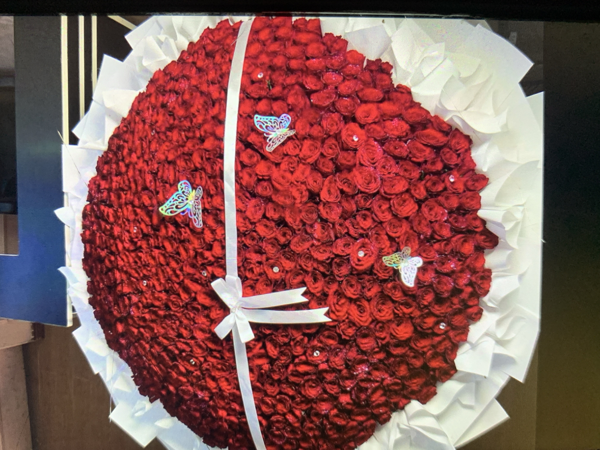 A large bouquet of red roses dominates the frame. The roses are densely packed in a circular or heart-shaped arrangement. A wide white ribbon is placed diagonally across the bouquet, tied in a bow near the center. Three decorative butterflies are attached to the roses. Small, transparent dew drops are scattered across the roses. The bouquet is surrounded by white tissue paper. The background suggests an indoor setting, possibly a florist shop or event space. There is no evidence of this image being in Rafin Par, Nigeria.