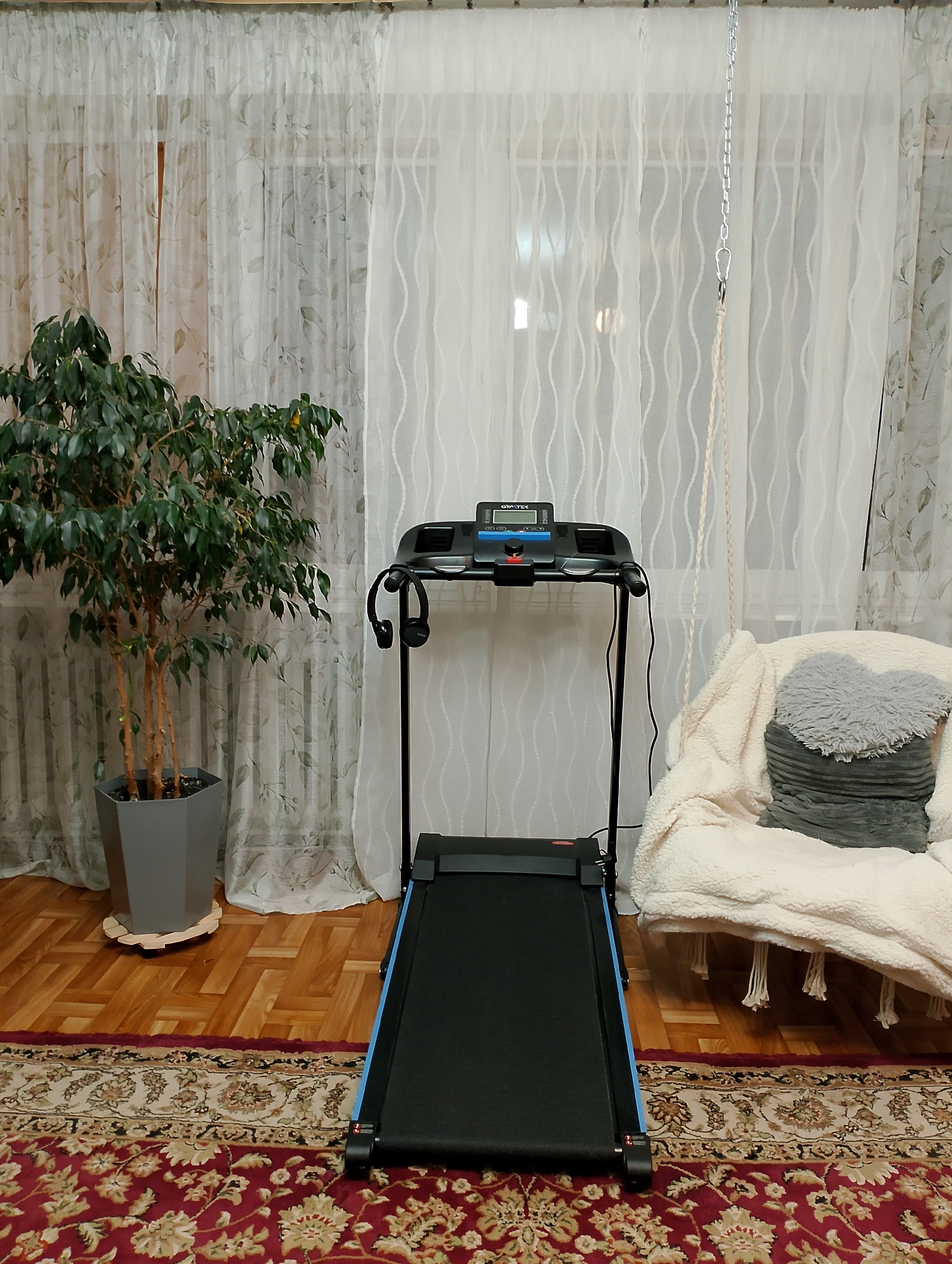 The image shows an indoor scene, likely a living room or exercise space, with a treadmill as the central focus. The treadmill is black with blue accents on the running belt and is positioned on a patterned red rug. To the left of the treadmill is a potted ficus tree in a hexagonal grey planter, which is elevated on a wooden base. In the background, sheer white curtains with a subtle floral pattern hang across a window. To the right of the treadmill, a plush, cream-colored armchair or beanbag chair is draped with a thick white blanket. A gray heart-shaped pillow with a ribbed texture rests on the blanket. A metal chain is suspended from the ceiling, connected to a braided rope that hangs down, suggesting the presence of a swing or hanging chair that is out of frame. The flooring is made of polished wooden planks arranged in a herringbone pattern. The overall impression is a home exercise setup combined with comfortable seating, suggesting a residential environment. There are no people visible in the image. The scene does not provide specific visual cues that would indicate its location in Kolno, Poland.