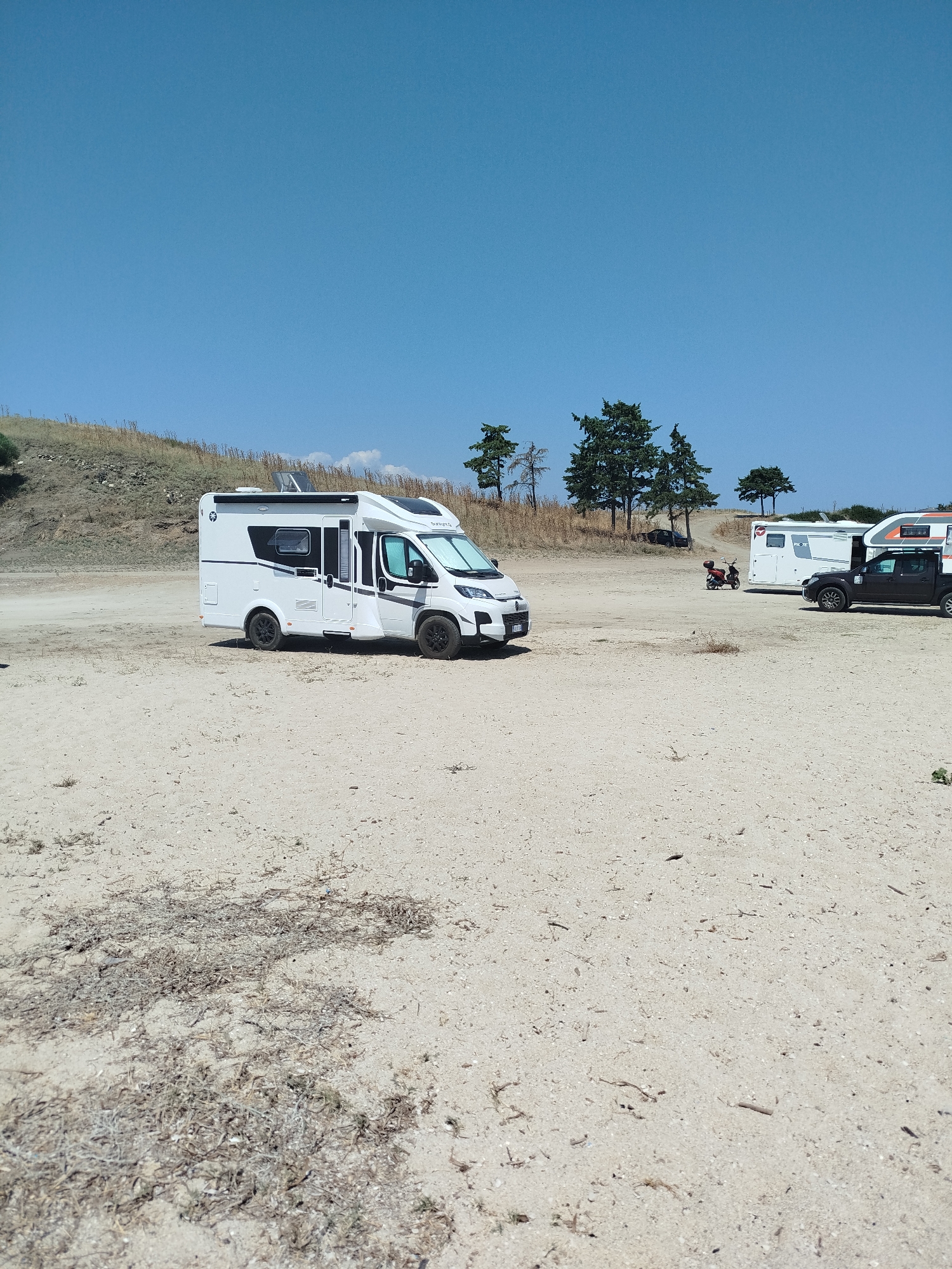 The image depicts a sandy area with several recreational vehicles parked. In the foreground, a white motorhome is prominently displayed, positioned slightly to the left of the frame. Further back, a white caravan, a red scooter, and a dark-colored pickup truck with a camper shell are visible. The background features a gentle slope covered in dry grass and scattered trees, under a clear blue sky. The scene appears to be a campsite or a parking area for recreational vehicles. The location is identified as Sithonia Municipal Unit, Greece.