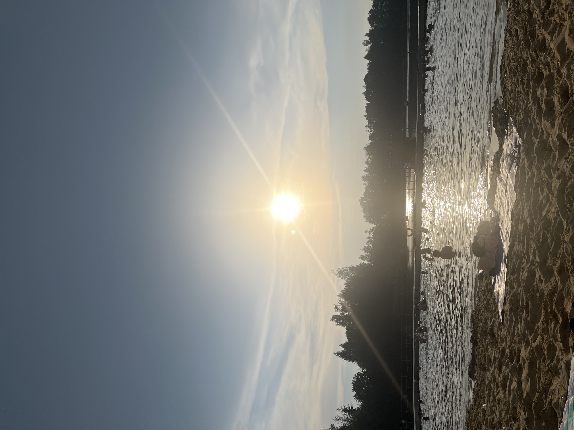 This is a vertical, wide shot of a beach on a sunny day. The sun is high in the sky and shining brightly, casting a glare on the water. The sky is a clear blue with a few wispy clouds. There are trees in the background, and a fence runs along the edge of the water. The beach is made of sand and there are a few people on it, some in the water and some on the sand. The water is calm and clear, and there are some ripples on the surface. The overall mood is peaceful and relaxing.