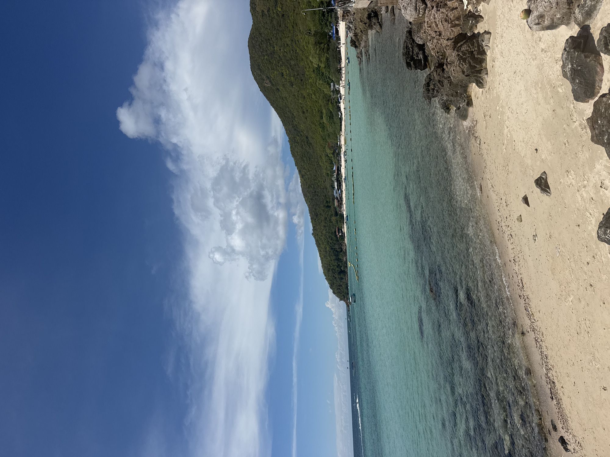 The image depicts a beach scene in Pattaya City, Thailand. A densely vegetated hill borders a sandy beach on the right, adjacent to the turquoise water. The shoreline features some rocky areas. The water is transparent, revealing the seabed. In the distance, along the beach, there are structures and people. The sky above is blue with scattered white clouds.