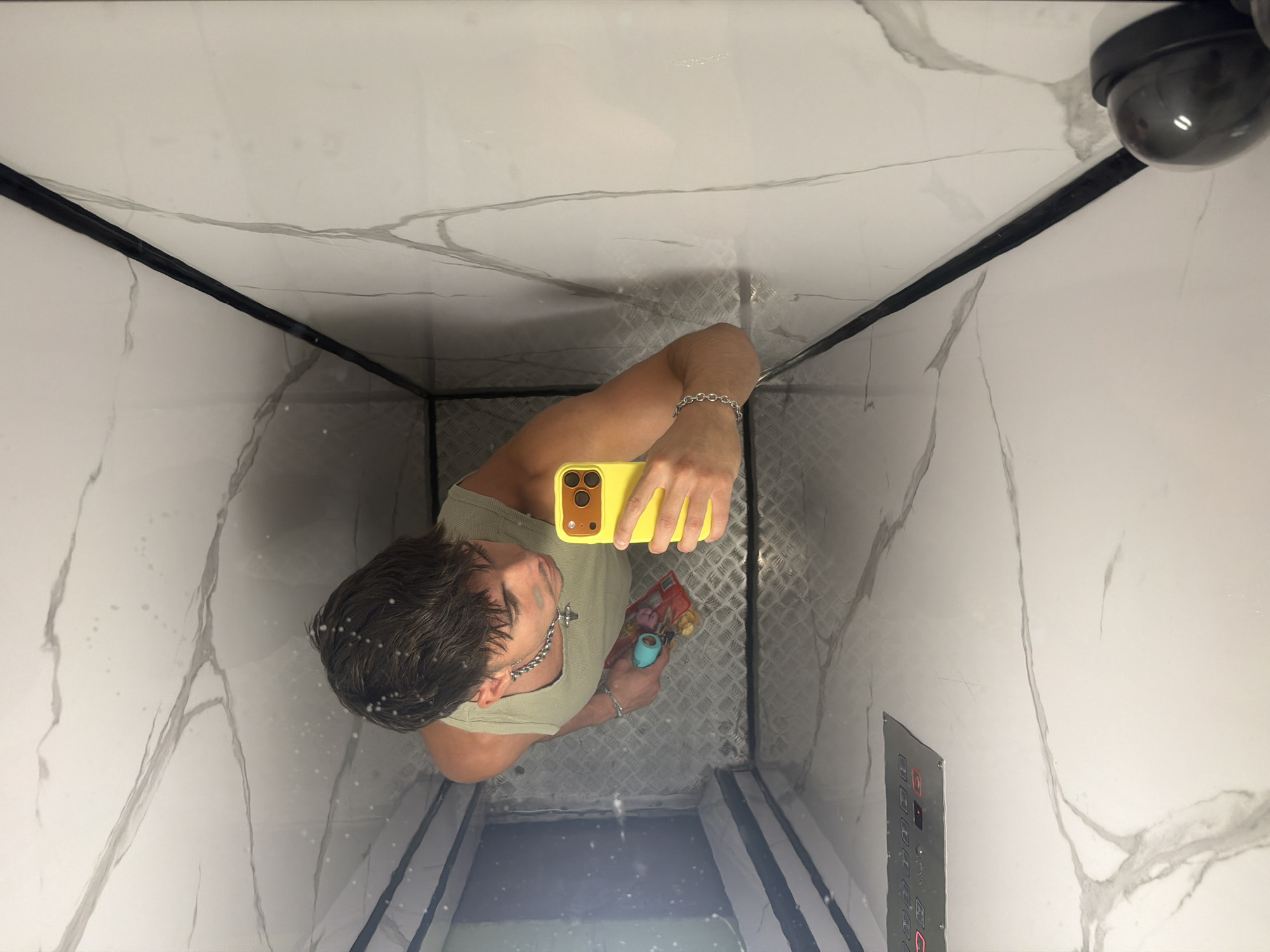 A young man with brown hair is taking a selfie in what appears to be an elevator. He is wearing a light green tank top and silver chain necklace, and a silver bracelet on his left wrist. In his right hand, he holds a yellow smartphone, and in his left hand, he holds a small object with a red wrapper and a blue handle. The elevator walls are covered in a white marble-patterned material with dark grey veins. The floor is a metallic, diamond-plate pattern. The lighting appears to be artificial, casting reflections on the shiny surfaces. The image is taken from a high angle, looking down at the man. The location is the city of Sofia, Bulgaria.
