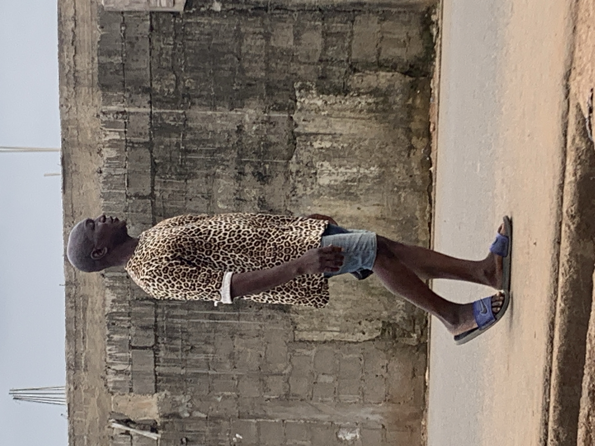 A photograph depicts a man standing in Akwanga, Nigeria. He is dark-skinned, bald, and wears a leopard-print top, short denim shorts, and blue sandals. The man stands on a concrete road next to a rough, gray, brick wall. A narrow path borders the road, leading to another textured wall. The sky appears overcast.