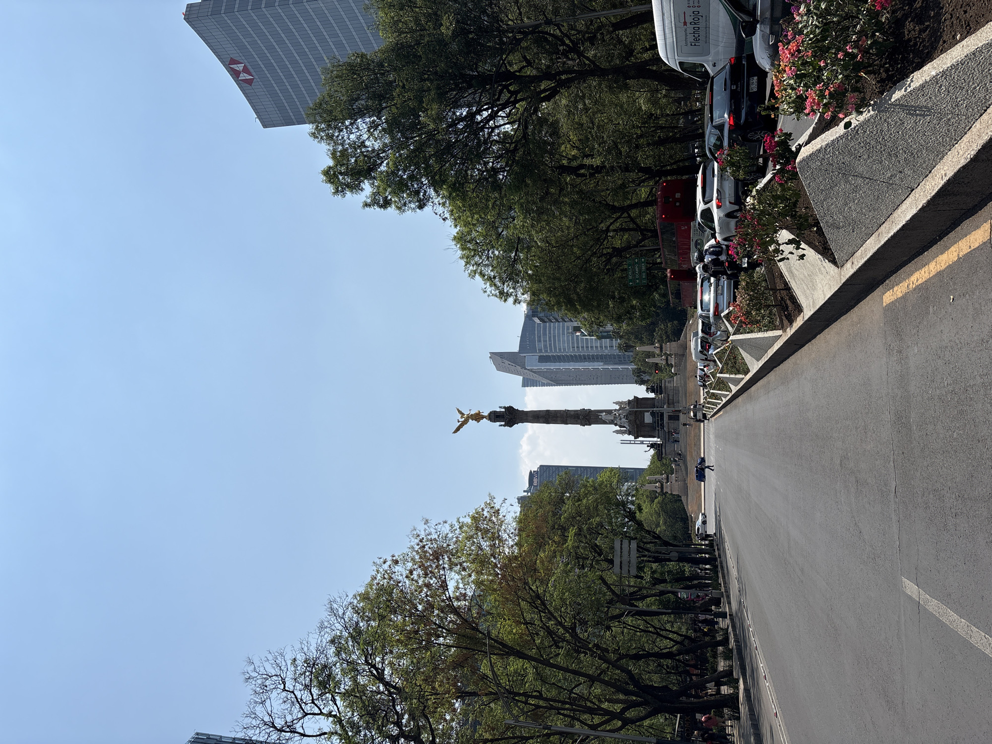 The image captures a street view in Mexico City, Mexico, featuring the iconic Angel of Independence monument in the distance. The roadway stretches into the distance towards the monument, lined with trees on both sides. A tall building with the HSBC logo is visible on the left side of the frame. The right side of the frame shows parked cars and a sidewalk with flowers. The sky is clear and blue. The scene appears to be during daylight. There are people walking on the streets and sidewalks.