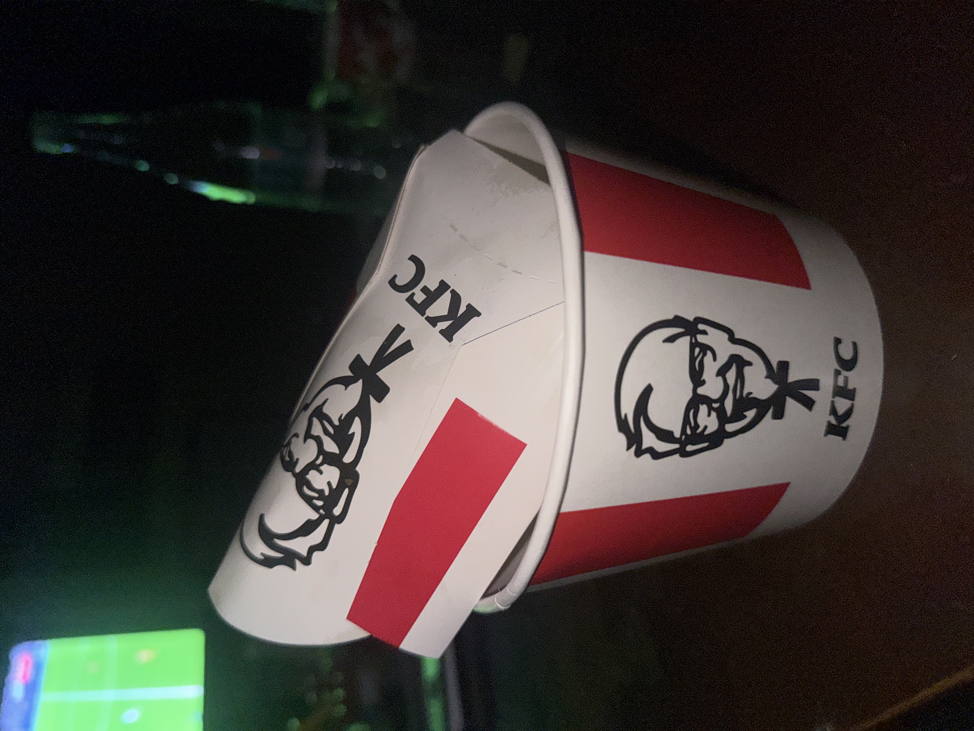 The image shows two stacked KFC buckets. The buckets are white with the KFC logo and red stripes. The buckets appear to be indoors, possibly on a table. In the background, there is a blurry green screen, likely a television. The location and city of Yerevan, Armenia cannot be definitively determined from the image.