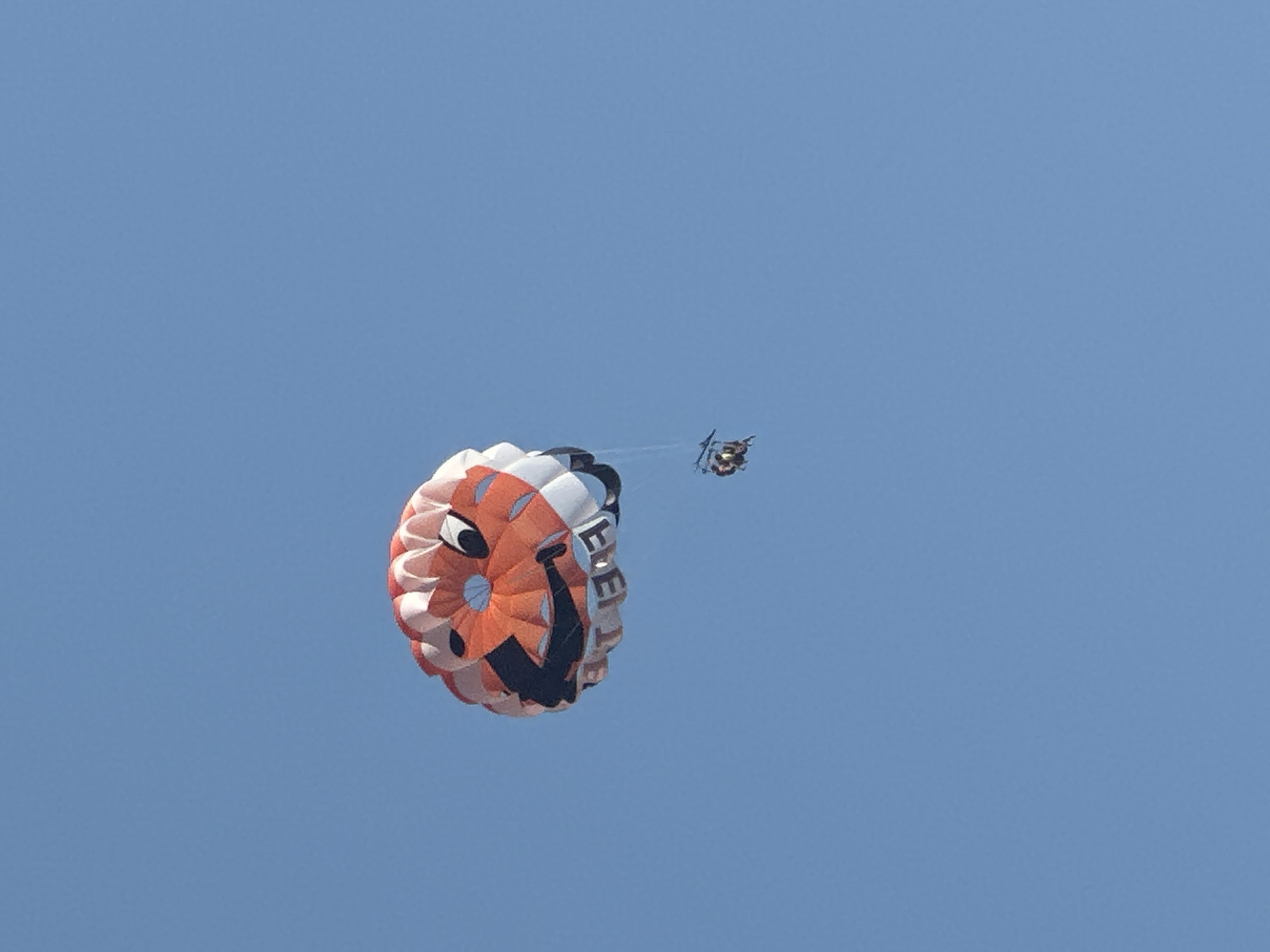 A parasail with a smiley face design is suspended in a clear blue sky. The parachute is predominantly orange and white, with a black smiley face featuring wide eyes and a broad grin. Text is visible along the bottom edge of the parachute, though it is partially obscured and difficult to read. Attached to the parachute by lines is a small passenger harness containing two individuals, appearing to be enjoying a recreational activity. The scene is observed from below, with the sky occupying the majority of the frame. There is no visible indication of the city of Boreti, Montenegro in this image.