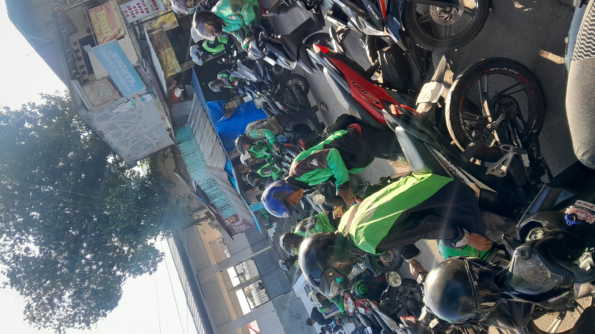 The image captures a street scene in Bandung City, Indonesia, likely during the daytime, characterized by a gathering of motorcycle riders. The riders are predominantly wearing green jackets, a common uniform for ride-sharing or delivery services, suggesting they are associated with platforms like Gojek or Grab. Several individuals are seated on their motorcycles, with some actively engaged with their mobile phones, possibly for work or communication.
The environment includes a street with asphalt paving, lined with buildings that appear to be commercial establishments, indicated by signage. One sign prominently displays "MIE KOKOK & BAKSO KARUNIA", identifying a food vendor. The background features a large tree with dense foliage, casting shadows and contributing to the bright, sunny atmosphere. Other motorcycles are parked or waiting, creating a sense of a busy hub or waiting area for these riders. The overall impression is a snapshot of a typical urban street environment in Indonesia, focusing on the daily activities of ride-sharing personnel.