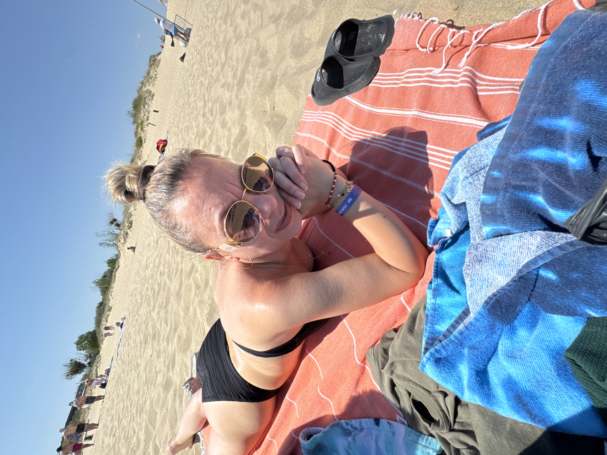 A woman sunbathes on a towel on a sandy beach. She is lying on her stomach, wearing a black bikini and sunglasses. In the background, other beachgoers are visible, as well as sand dunes and the sky. The scene is located on the beach in Nesebar, Bulgaria.
