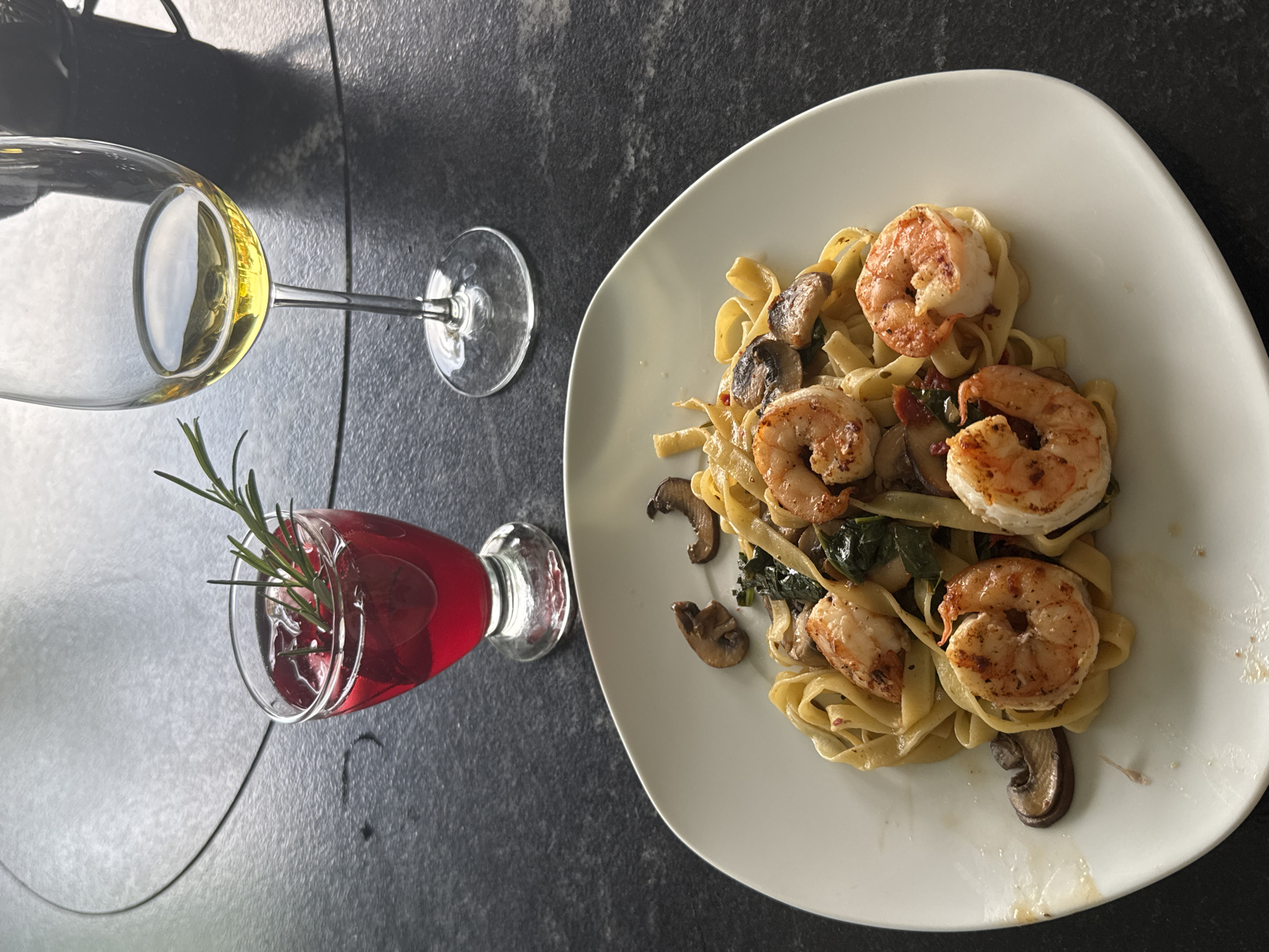 The image shows a place setting on a dark, textured surface resembling stone or concrete. A white plate with tagliatelle pasta topped with shrimp, mushrooms, and possibly spinach or other greens is placed on the right. To the left of the plate are two stemmed glasses; one contains a light yellow liquid and the other a red liquid with a sprig of rosemary in it. The scene is indoors, presumably on a table. There are no identifiable landmarks or people to verify the location is San Bernardino Tlaxcalancingo, Mexico.