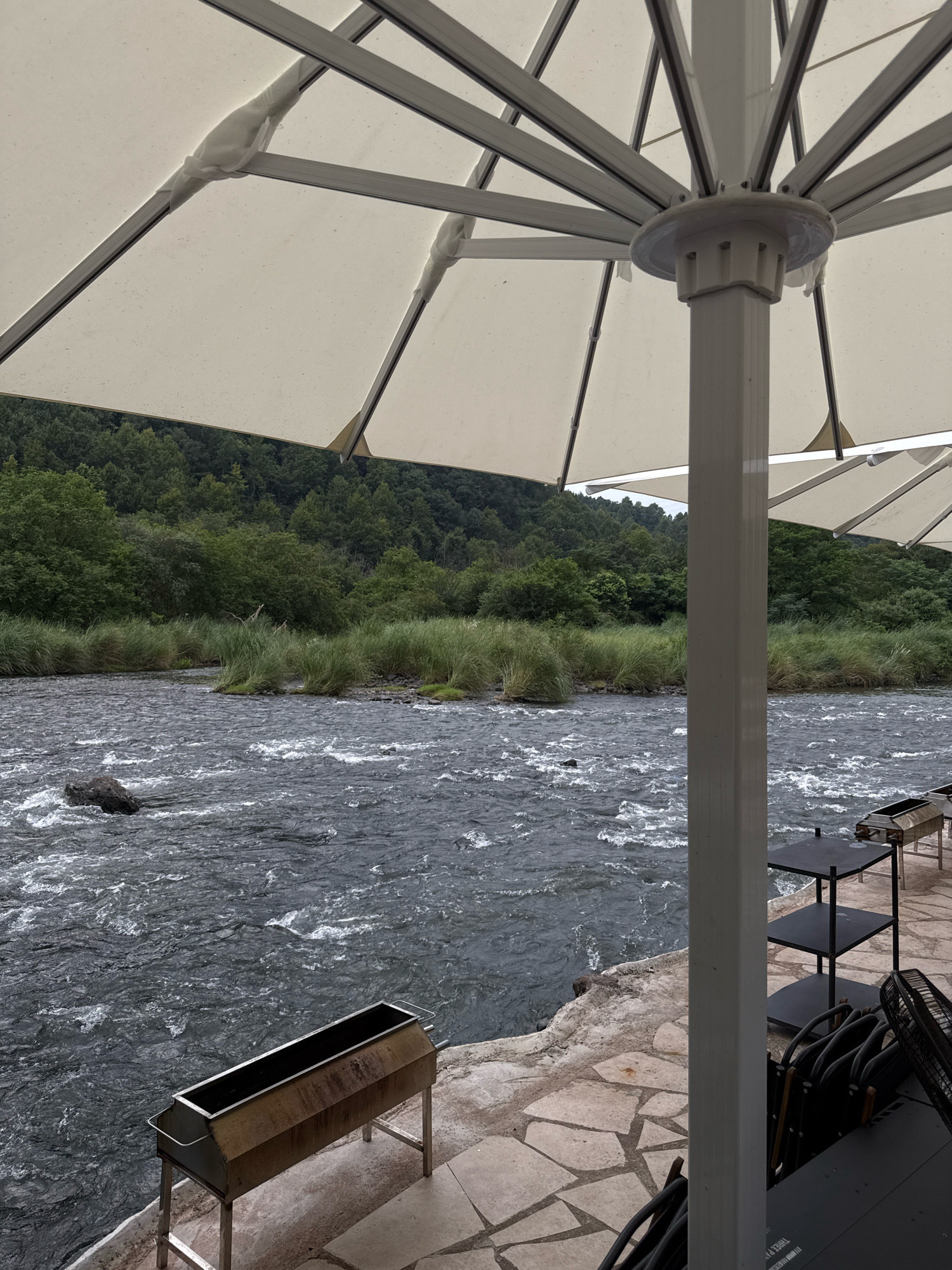 The image depicts an outdoor seating area next to a flowing river. A large, cream-colored umbrella with a white central pole provides shade. In the foreground, there are two stainless steel barbecue grills. To the right, black tables and stacked chairs are visible. The river is dark and choppy, with a large rock in its center. The riverbank is lined with tall, green reeds and grassy areas, beyond which is a dense forest of green trees covering rolling hills. The ground in the seating area is paved with irregular flagstones. The scene is tranquil and appears to be a riverside dining or recreational space.