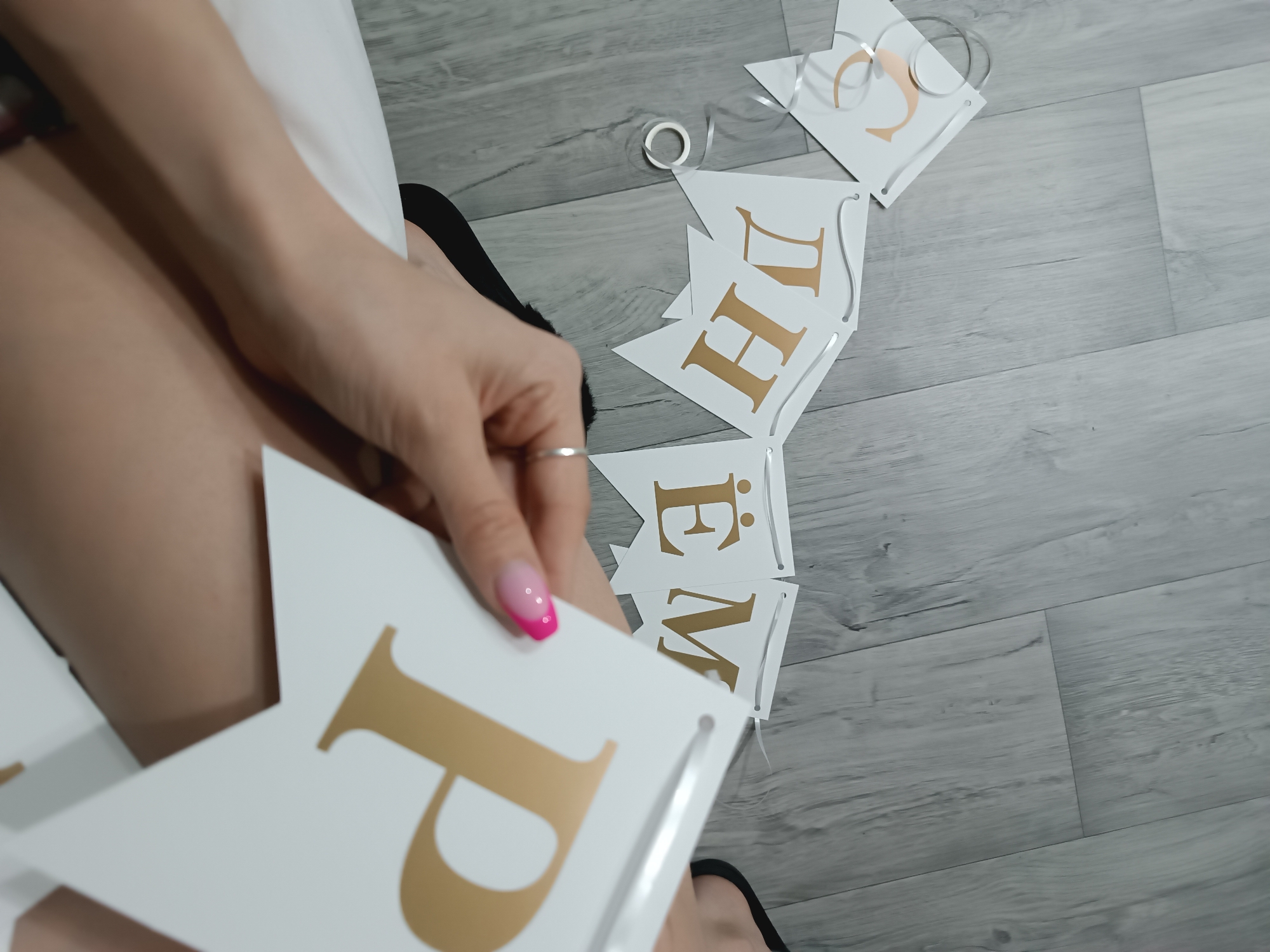 The image depicts an indoor scene at an unspecified location in the city of Unknown, Unknown. A person is observed from an overhead perspective, with their bare legs, clad in a white garment, and right hand prominently in the foreground. The person's right hand, adorned with a silver ring on the ring finger and featuring a pink-to-clear ombré manicure, holds a white triangular banner flag with a gold Cyrillic letter "Р" (R). A black slipper or fuzzy slide is partially visible on the person's left foot. Scattered on the light grey wood-look laminate floor are additional white triangular banner flags with gold Cyrillic letters, including "О", "Ж", "Д", "Н", "Ё", "М", and "С", forming parts of the phrase "С ДНЁМ РОЖДЕНИЯ" (Happy Birthday). The flags are connected by silver ribbon, and a spool of silver ribbon is also present on the floor. The environment consists of horizontally laid planks of light grey flooring.
