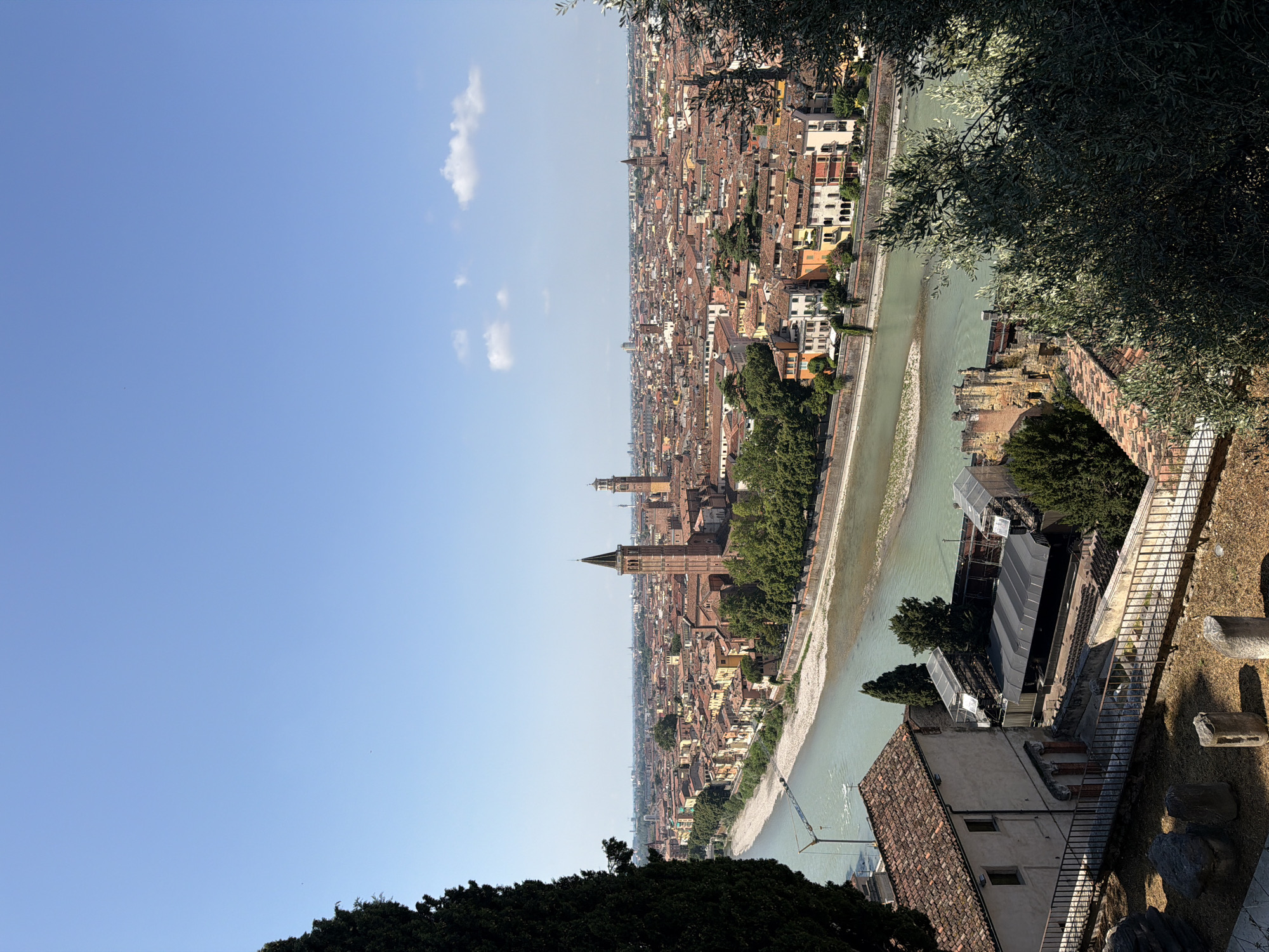 This is a high-angle, wide-shot photograph of Verona, Italy, on a clear, sunny day. The cityscape is dominated by terracotta-tiled roofs and the Adige River, which bisects the city. In the foreground, a large, dark green evergreen tree partially obscures the view on the left. To the right and foreground, olive branches are visible.
On the left side of the image, in the mid-ground, a building with a terracotta roof is situated near the riverbank. Adjacent to it, an outdoor stage area is set up with a dark covering and scaffolding. In front of this structure, a low metal railing follows a path or walkway. Scattered on the ground in this area are several large, rough-hewn stones and two cylindrical stone columns, suggesting historical ruins.
The Adige River flows from left to right across the image, appearing a murky green. Along its banks, stone walls and trees are visible. Across the river, the dense urban fabric of Verona unfolds. Numerous buildings, primarily with red-tiled roofs, fill the landscape. Two prominent church towers rise above the rooftops, one taller and more slender with a pointed spire, and another shorter and wider with a dome-like top. The distant horizon is hazy, with hints of more buildings and possibly hills. The sky is a bright, cloudless blue. No people are visible in the photograph.