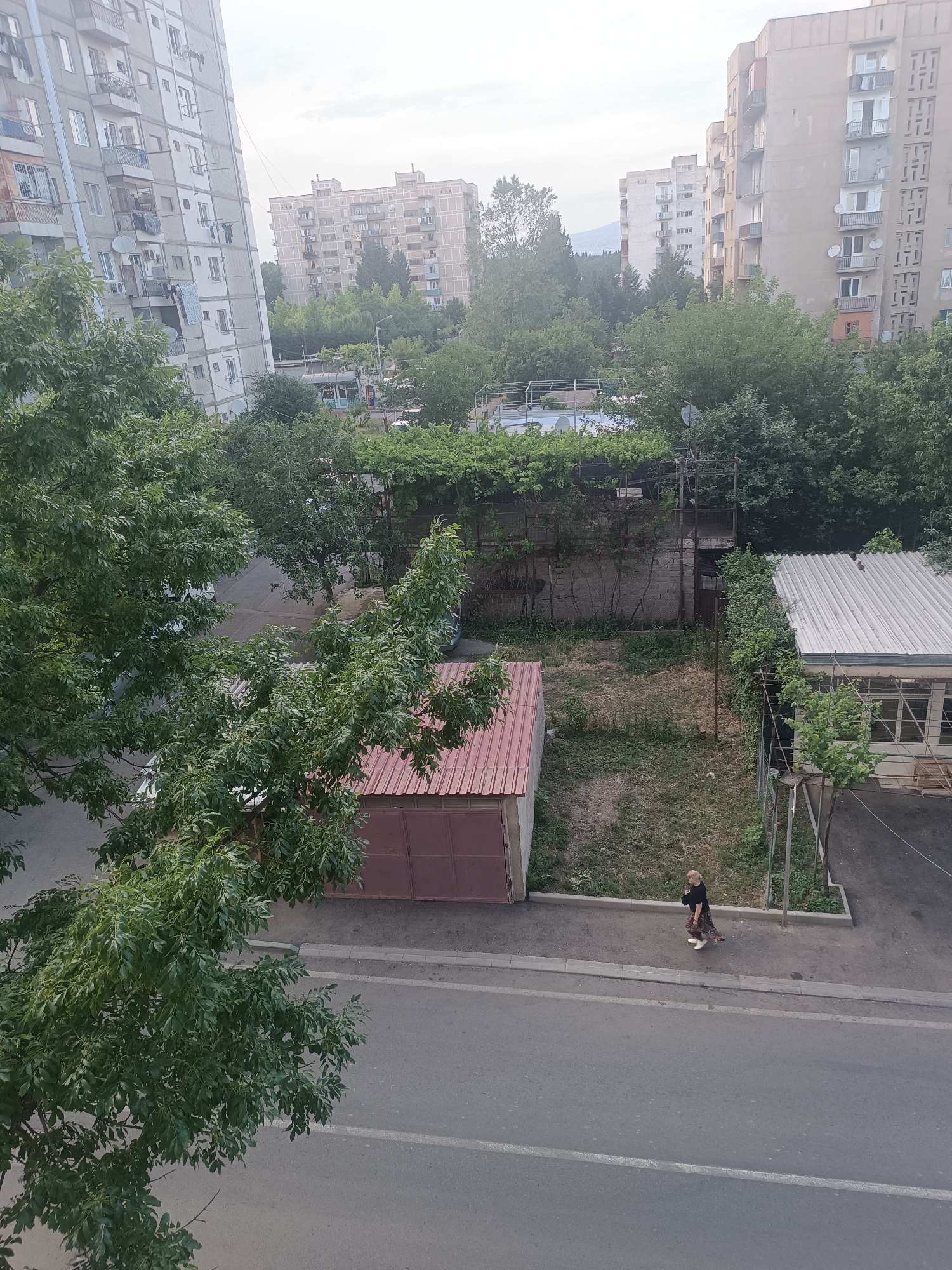 This media file captures an outdoor scene in Tbilisi, Georgia. The setting appears to be a residential area with several tall apartment buildings visible in the background, characteristic of Soviet-era architecture. Lush green trees are abundant, some with dense foliage, suggesting a warm season.
In the foreground, a paved road runs horizontally across the frame, with a sidewalk bordering it. A lone person, a woman with blonde hair, is walking on the sidewalk from right to left. She is wearing a dark top and a patterned skirt, with light-colored shoes. She is carrying a white object in her left hand, possibly a bag or a small dog.
Behind the sidewalk, there are several structures. A small, single-story building with a red, corrugated metal roof is visible on the left. Further back, there are fences, more greenery, and the foundations of other buildings. In the distance, more apartment blocks and trees create a layered urban-meets-nature landscape.
The sky is overcast, indicating a cloudy day. The lighting suggests it is either early morning or late afternoon, with diffused light. No visible text is present in the image. The overall impression is a quiet, everyday moment in a residential part of the city.