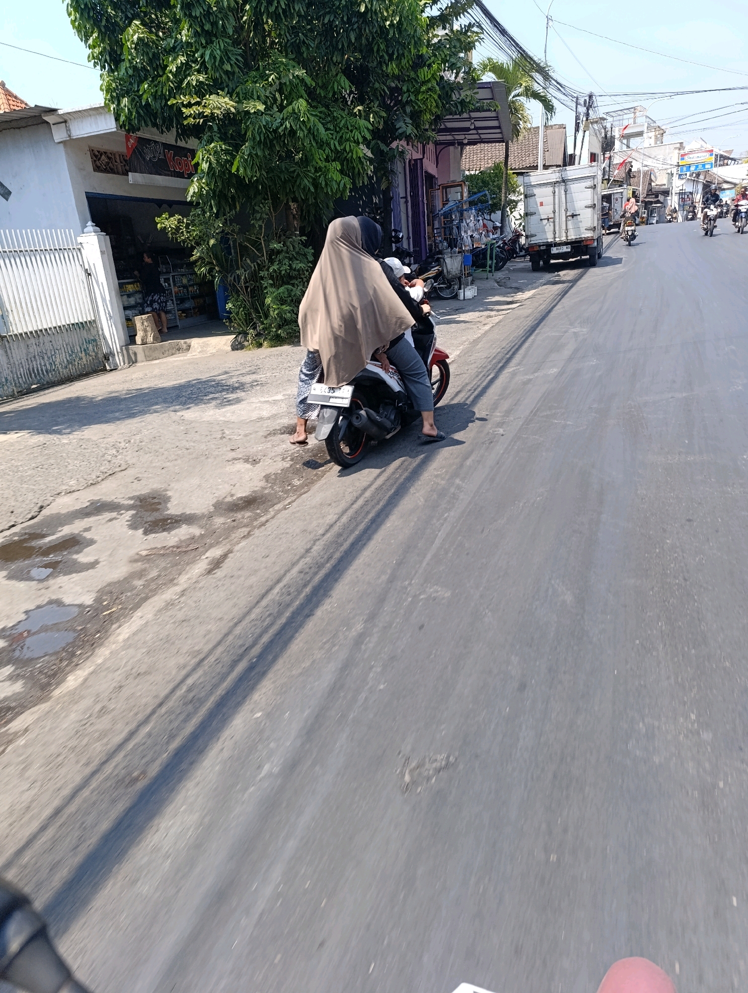 The image shows a street scene in Glagah Sari, Indonesia. In the foreground, two people are on a red and white motorcycle, with the rider wearing a brown hijab and gray pants, and the passenger also wearing a brown hijab. They are on a paved road with some patches of wetness and debris. To the left, a white building with a metal fence and a sign that says "KOPI" is visible. A person is standing outside the building. Further down the street, a white truck is parked, and several other motorcycles and people are visible. The environment appears to be a commercial area with shops and buildings lining the street. The sky is clear and blue, indicating a sunny day.