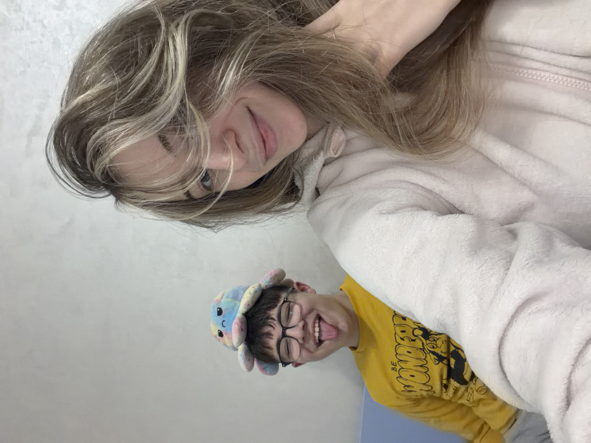 Two individuals are pictured indoors. The person in the upper portion of the frame is a young woman with long blonde hair. She is wearing a light-colored, possibly pink, fuzzy jacket. The person in the lower portion of the frame is wearing glasses, a yellow t-shirt with black lettering and a cartoon graphic, and a novelty hat resembling an octopus. The background appears to be a plain wall, possibly in a residential setting. The city of Plovdiv, Bulgaria, is not discernible from the visual details of the image.