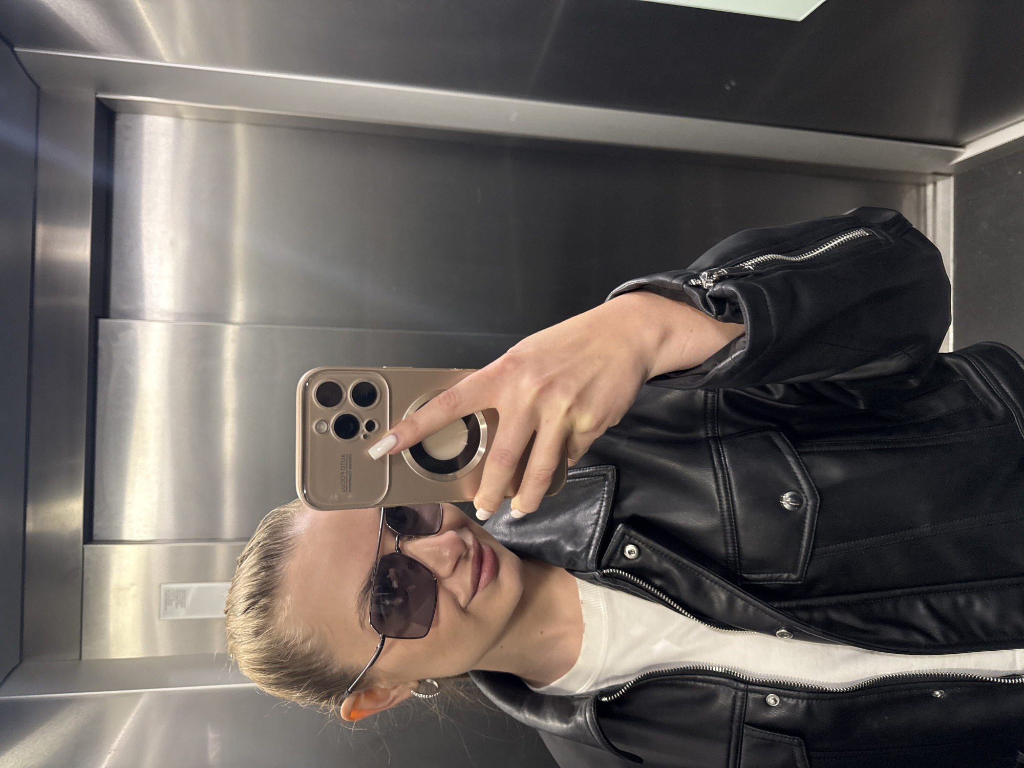 A woman is taking a selfie in what appears to be an elevator. She is wearing a black leather jacket, sunglasses, and a white shirt underneath. The elevator walls are made of brushed metal. The phone she is using is a gold-colored iPhone. The location is possibly in Budva, Montenegro, but this cannot be confirmed solely from the image. The subject is holding her phone up to take the picture.