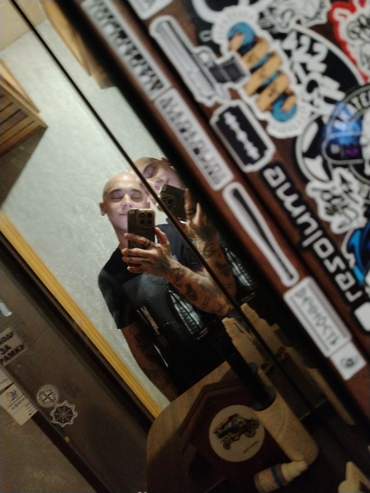 A mirror reflects a bald man holding a smartphone, taking a selfie. The man is wearing a dark t-shirt and has tattoos on his arms. He is looking at his phone and appears to be smiling. The background in the reflection shows a wall with a subtle pattern. On the surface of the mirror, or an object adjacent to it, there are various stickers with designs and text. One sticker appears to be a razor blade. Another sticker has text that reads "resulum." In the foreground, a wooden frame is visible. To the left of the frame, a door or window has some text printed on it, possibly in Cyrillic script, along with two circular stickers. One sticker features a compass-like design, and the other is a spiderweb design. Further down, another sticker with a vehicle is visible. The overall scene appears to be indoors, possibly in a room or hallway.
