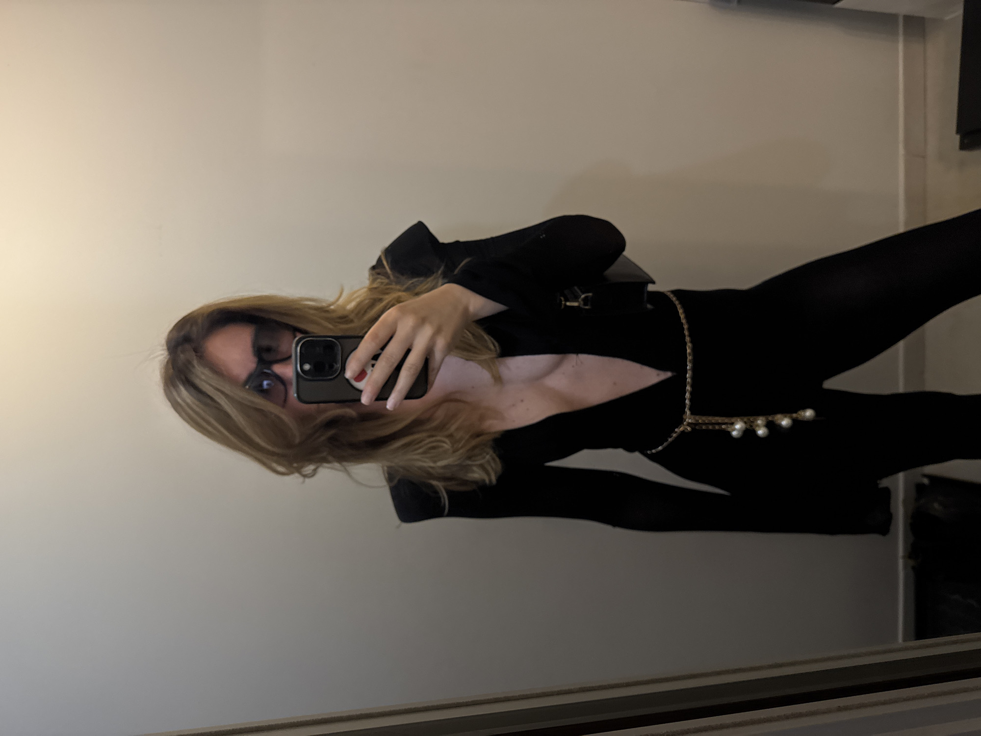 A woman with blonde hair and sunglasses takes a mirror selfie in a room with a light-colored wall. She is wearing a black jumpsuit with a plunging neckline and a gold chain belt with pearls. The room appears to be a domestic setting, possibly a bedroom or dressing room, with a mirror on the left side of the frame reflecting a dark object on the floor. There is a gray object on the right side of the frame. The location is not specified in Sofia, Bulgaria.